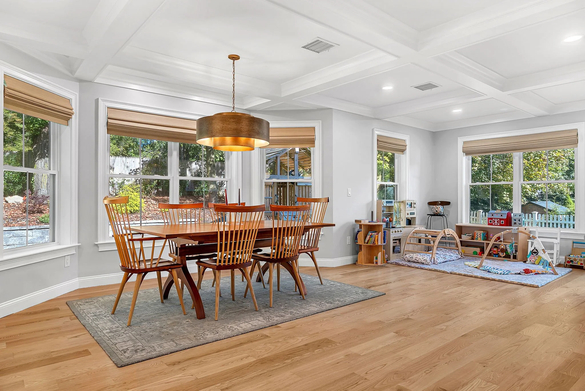 A spacious dining and playroom with a wooden dining table and eight chairs on a gray rug, large windows with beige blinds, a wooden chandelier, and a play area with toys, books, and a play mat for children.