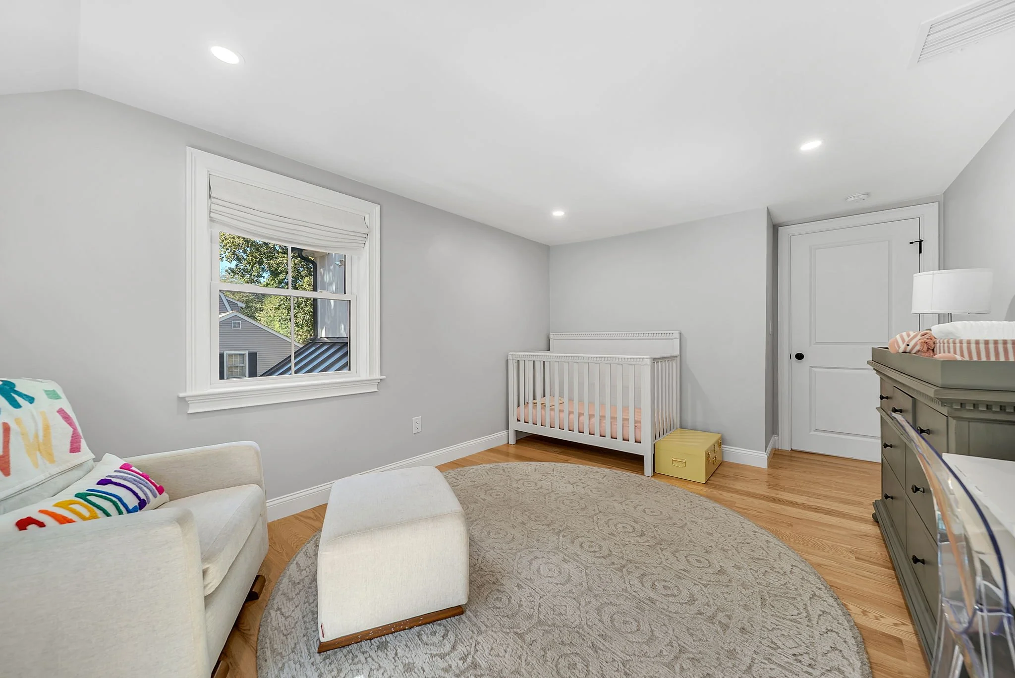A nursery room with white walls, hardwood floors, a window with white trim, a white armchair with colorful pillows, a matching ottoman, a white crib with pink bedding, a yellow storage box, and a gray dresser with a lamp. The room is lit by ceiling l