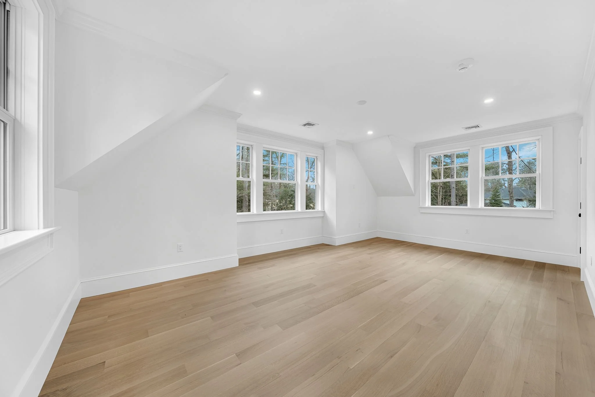 Empty room with white walls, multiple windows, light wood flooring, and ceiling lights.