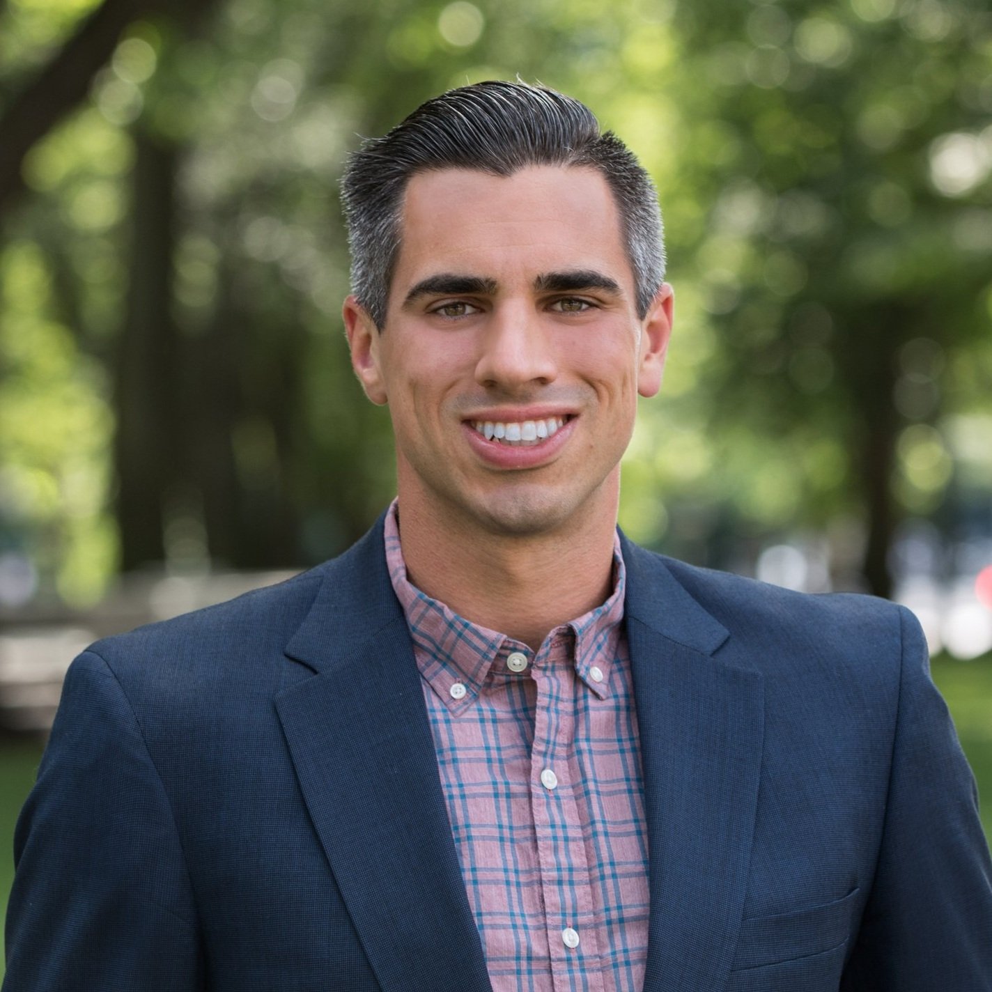Portrait of a smiling person in a blue blazer and checkered shirt, standing outdoors with a blurred green background.