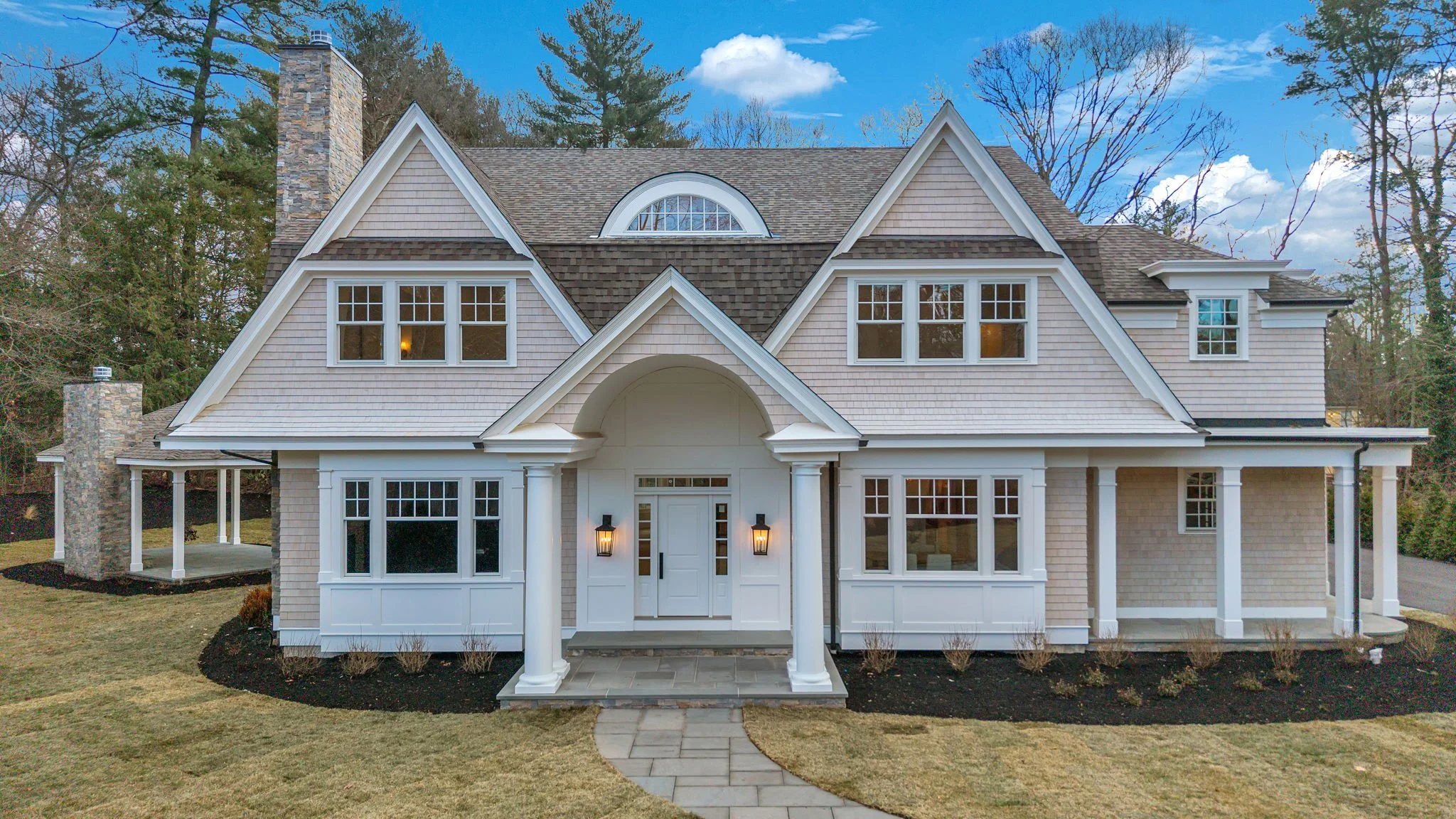 Front view of a large, modern house with white walls, multiple windows, a covered porch, and a sloped roof surrounded by a landscaped yard with grass and small bushes.