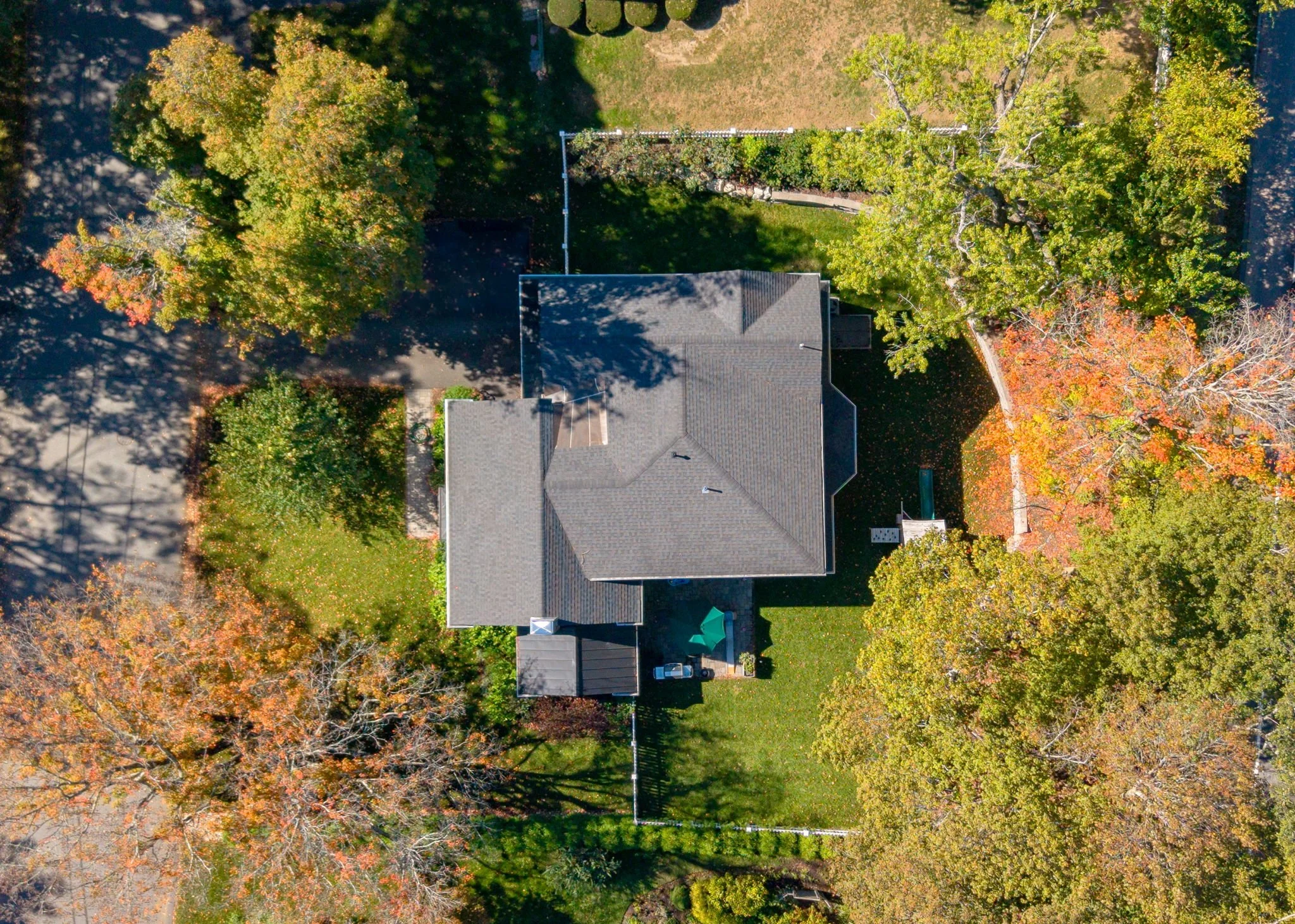 An aerial view of a house surrounded by trees with fall foliage, a green lawn, and a driveway.
