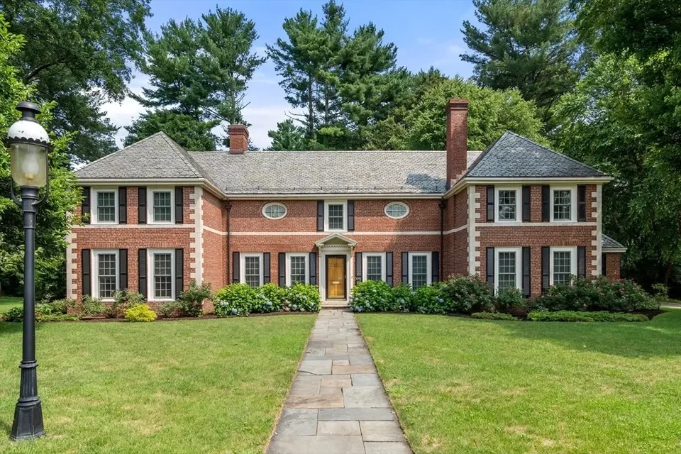 Large brick house with multiple windows, a front porch, and well-maintained lawn surrounded by trees.