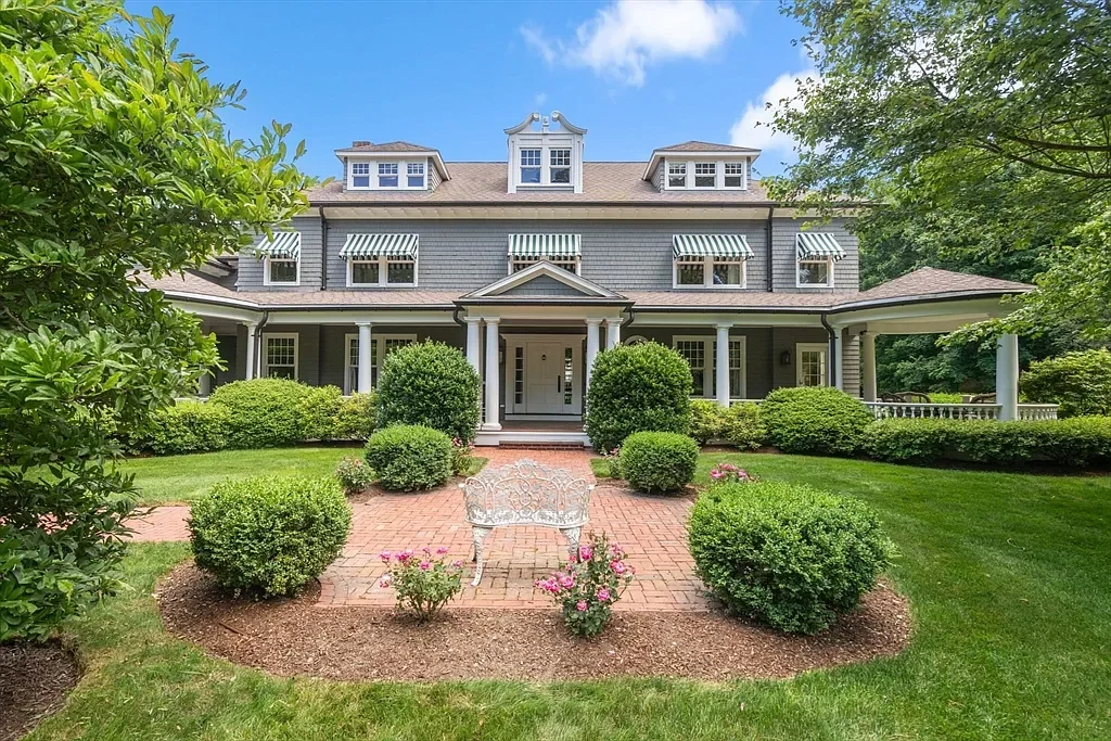 Front yard of a large, gray, two-story house with a covered porch, landscaped garden with bushes, flowers, and a brick pathway with a metal bench in the center.