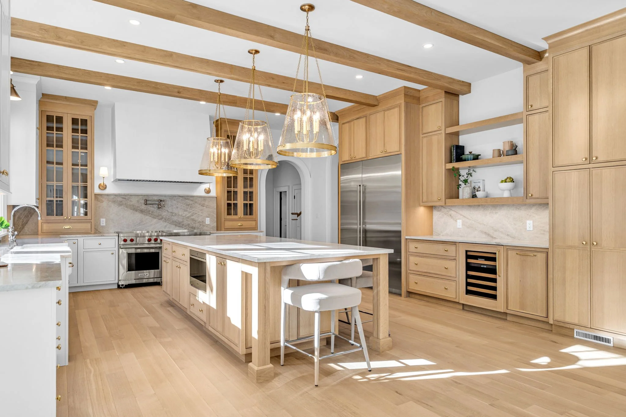 A spacious kitchen with a large central island, wooden cabinetry, and pendant lighting. There is a stainless steel refrigerator, a stove, open shelves with decor, and large windows allowing natural light.