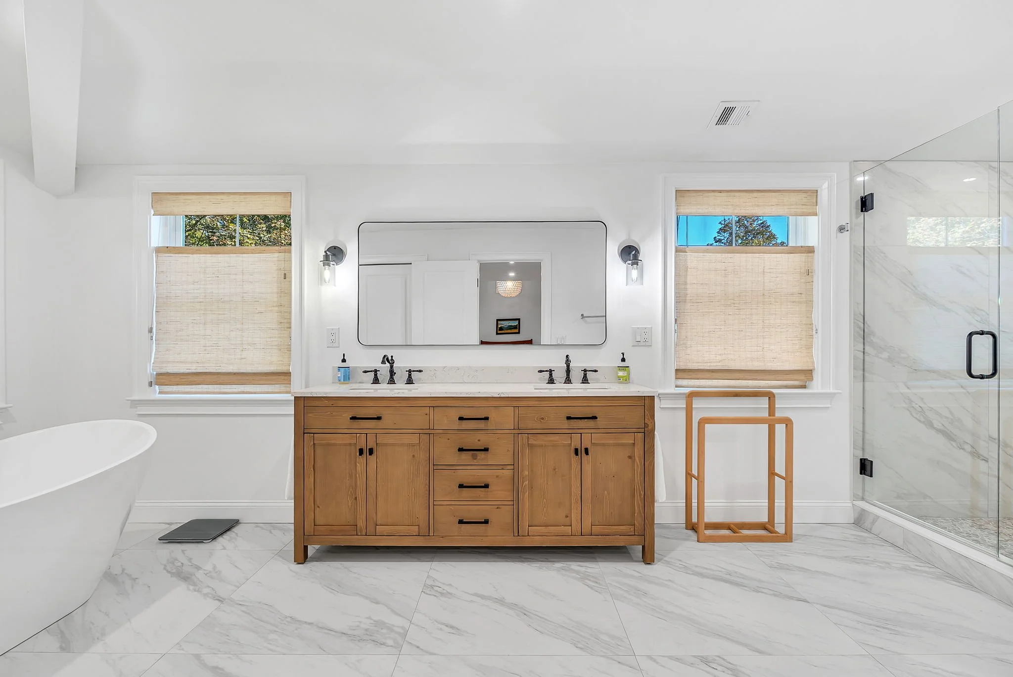 Modern bathroom with a wooden vanity, large mirror, two windows with bamboo shades, a white bathtub, and a glass shower enclosure with marble tiles.