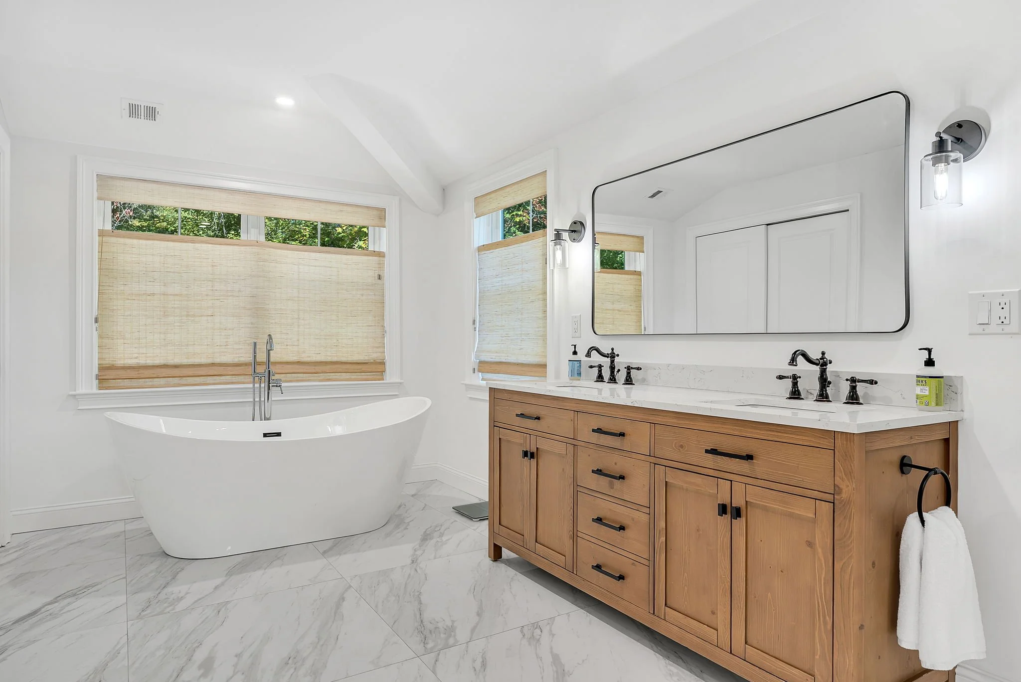 Modern bathroom with white walls, large mirror, wooden vanity with black hardware, corner bathtub with windows in the background, marble flooring, and soft natural lighting.