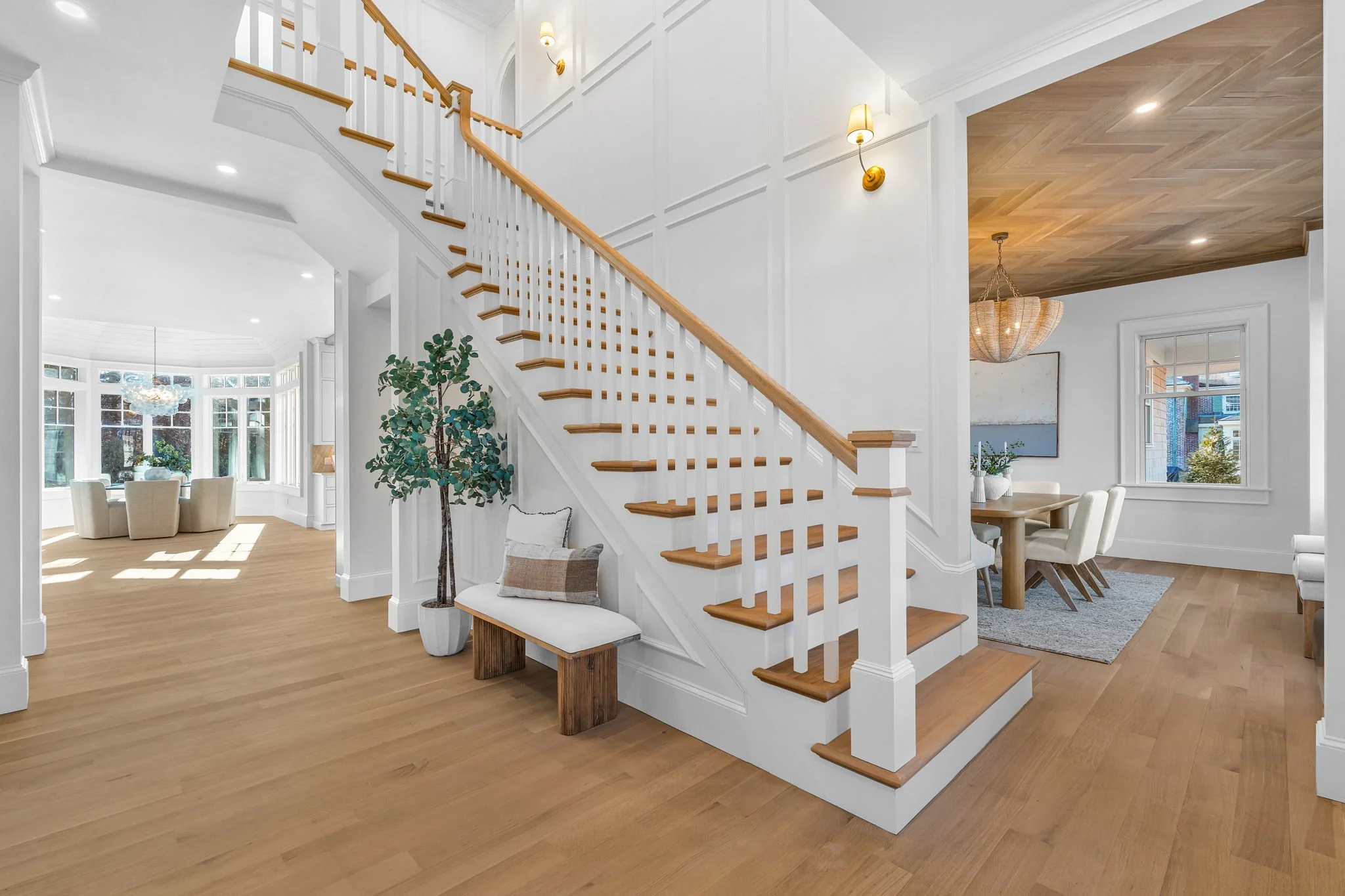 Interior of a modern home featuring a staircase with white spindles and wooden handrail, a sitting area with a bench and decorative pillows, and a dining room with a light wooden table and white chairs, all illuminated by natural light from large win