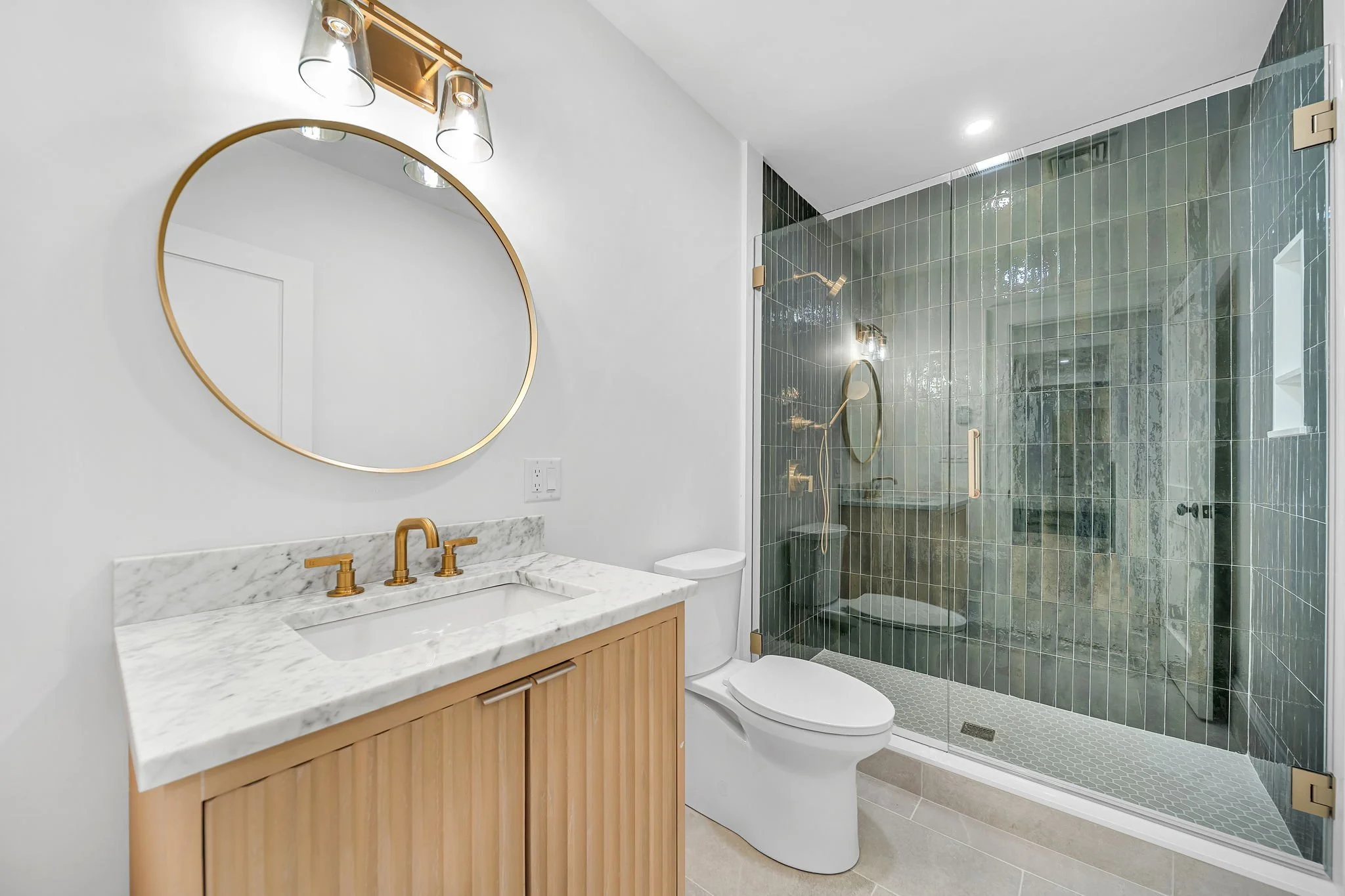 Modern bathroom with white walls, a marble countertop vanity with gold fixtures, a round mirror, a toilet, and a glass-enclosed shower with green tiles and gold accents.