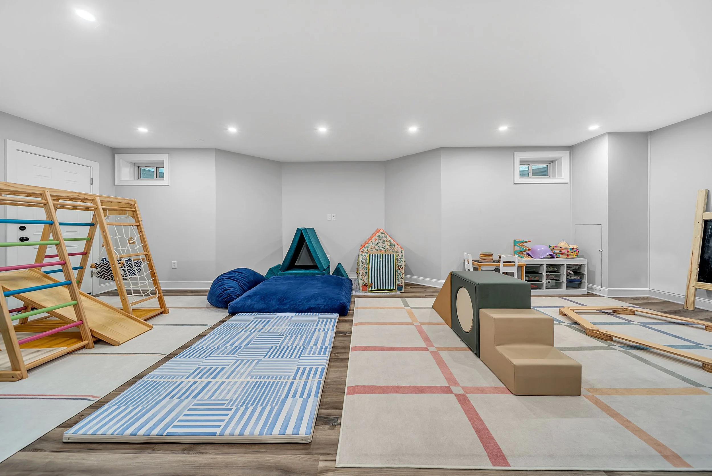 Indoor children's playroom with mats, soft play equipment, climbing structures, and storage shelves on a light gray wall background.