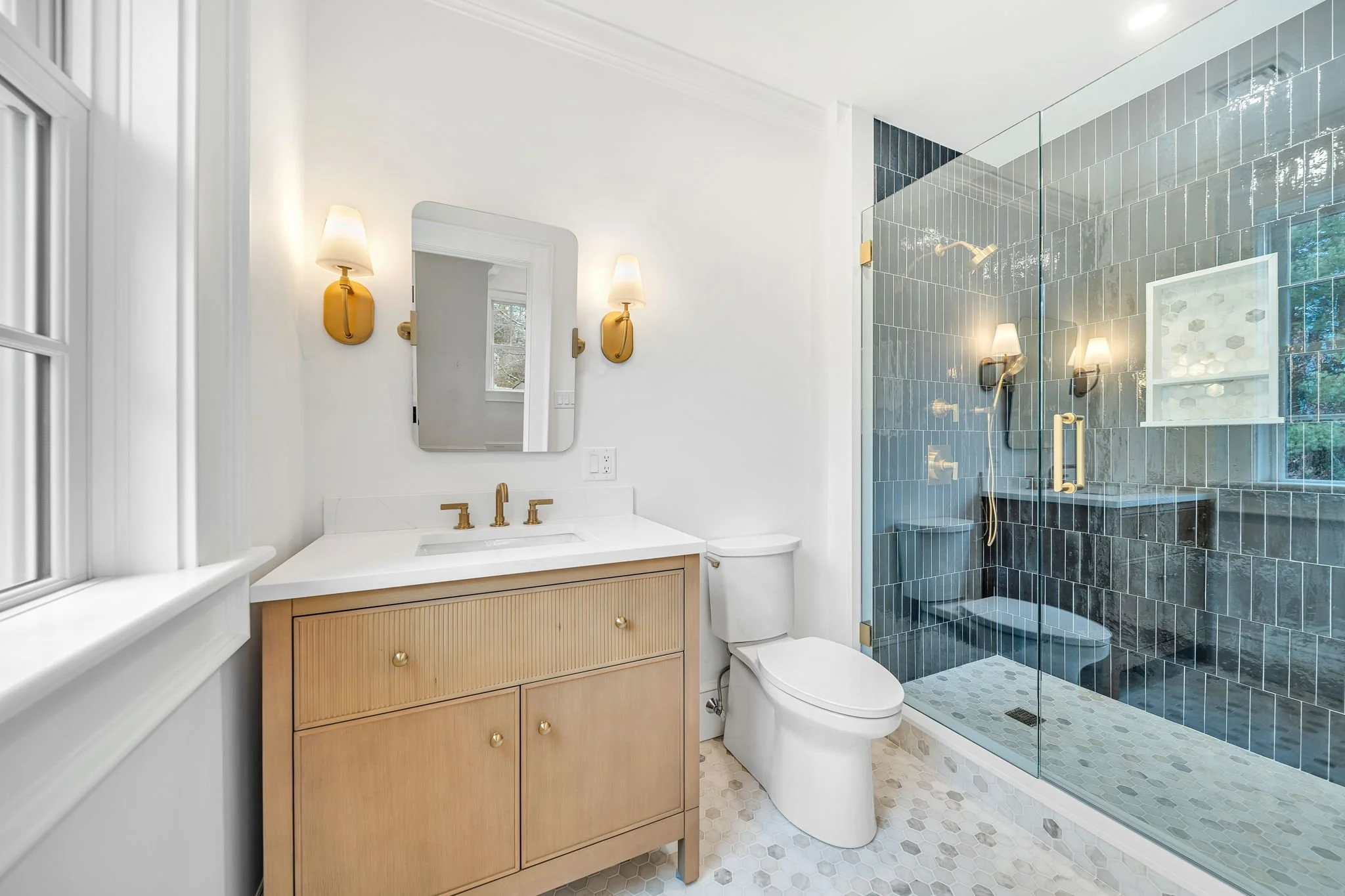 Bathroom with a wooden vanity, white countertop, gold fixtures, and a mirror. Next to it is a white toilet. The shower area has dark tiles, gold fixtures, a glass door, and windows with natural light.