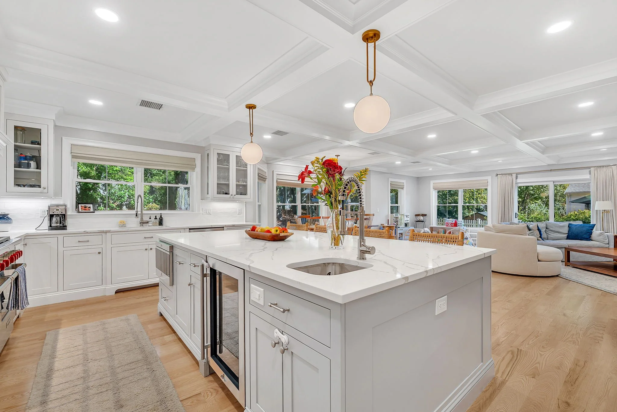 Spacious, modern kitchen with white cabinetry, marble countertop island, and large windows overlooking a lush green backyard.