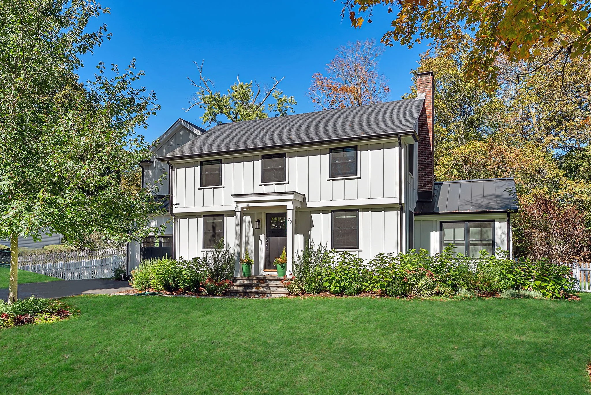 Front view of a two-story white house with black window frames, a small porch with stairs, surrounded by green lawn and bushes, under a clear blue sky with some trees in fall colors.