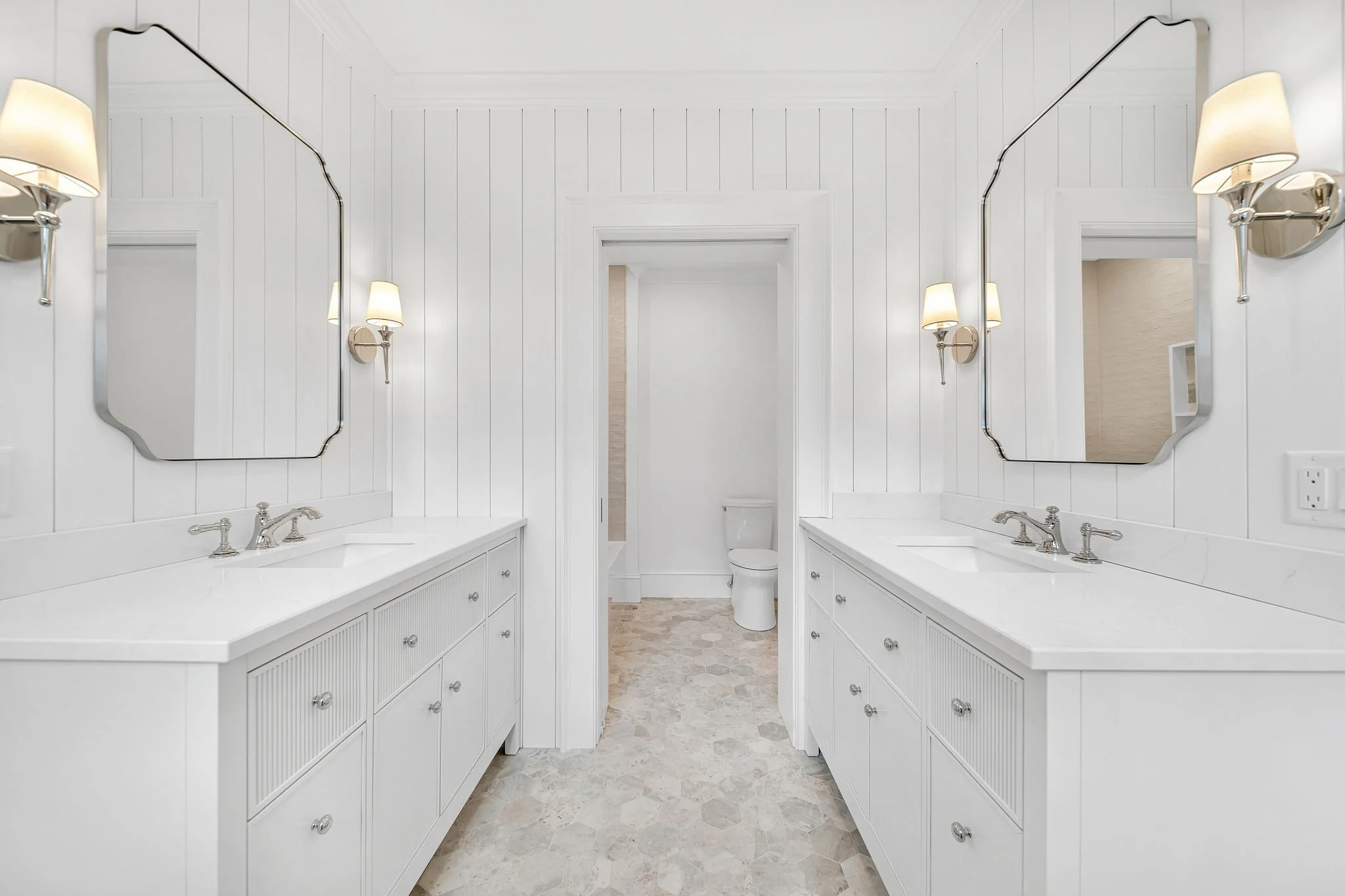 White bathroom with double vanities, large mirrors, wall-mounted lights, and a toilet in the background.