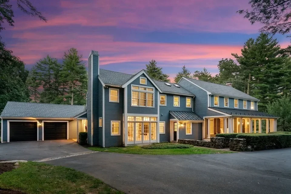 A large blue house with multiple windows illuminated from inside, surrounded by trees and a driveway at dusk.