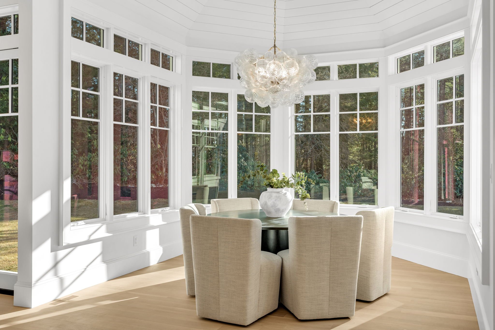 Sunlit octagonal dining room with large front windows, cream-colored chairs around a round table, and a modern chandelier hanging from the ceiling.