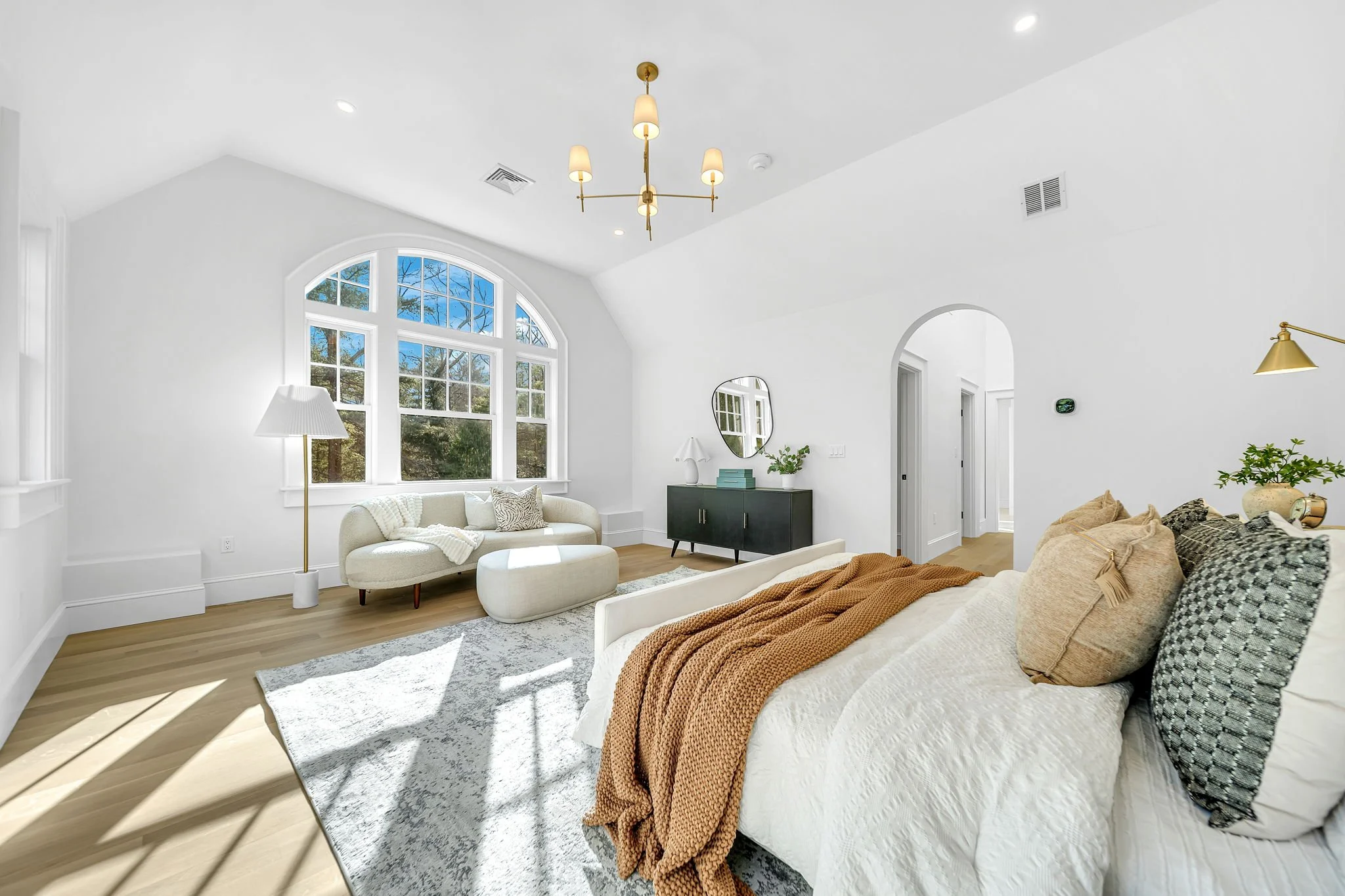 Bright living room with large arched window, white walls, beige sofa with pillows, white armchair with throw blankets, wooden flooring, modern chandelier, black sideboard with mirror and plants, open doorway leading to hall.