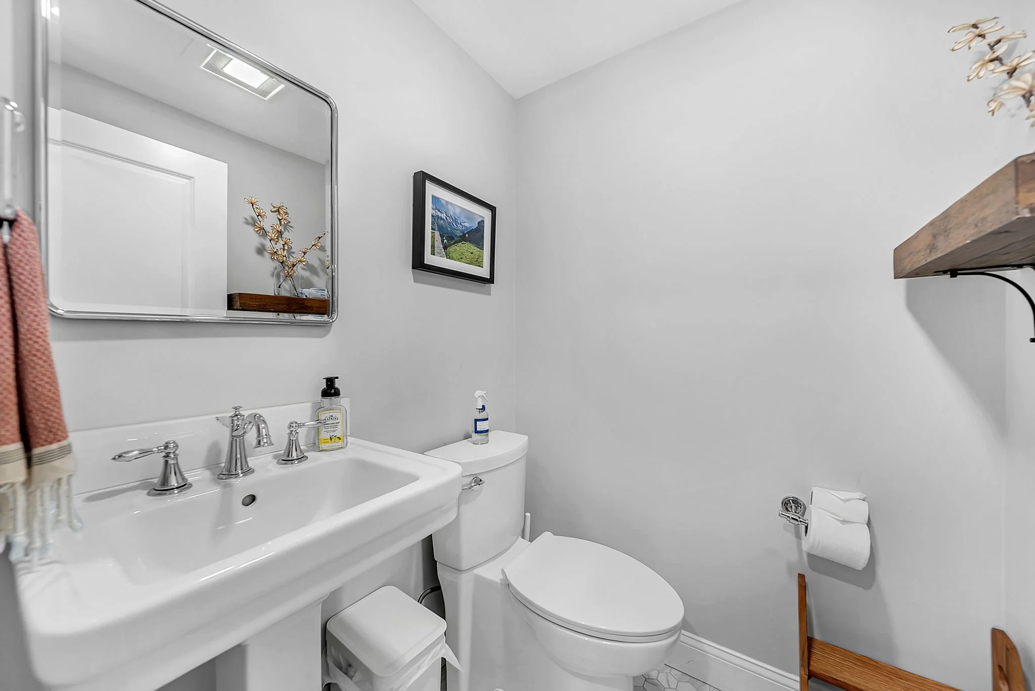 A small white bathroom with a wall mirror above a porcelain sink, a toilet, a wall-mounted picture of mountains and greenery, a spray bottle, a roll of toilet paper on a holder, and a wooden shelf on black brackets.