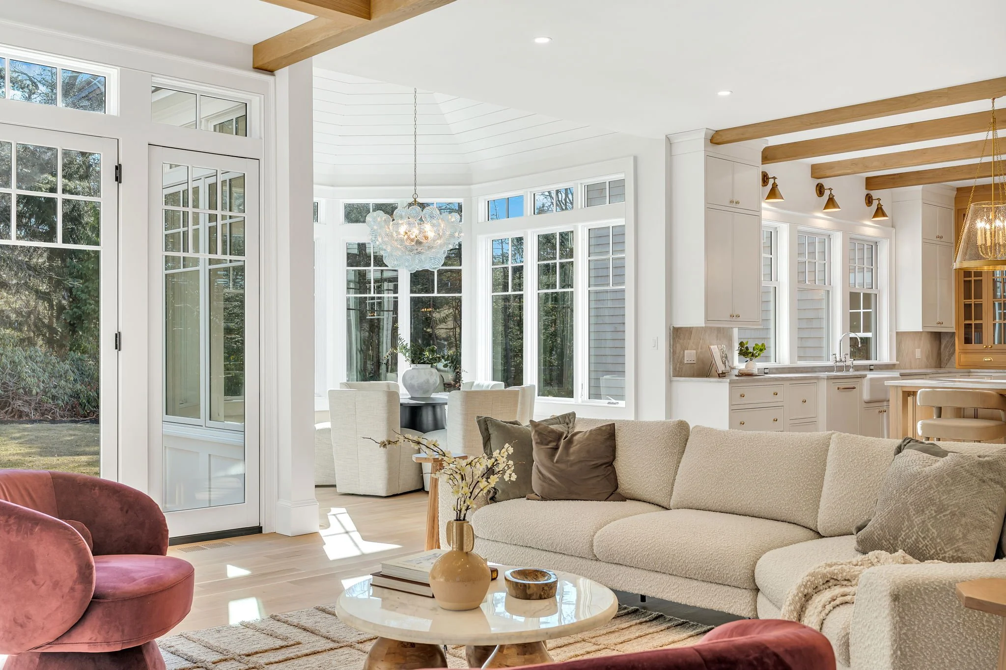 Bright, airy living room with a beige sofa, pink velvet chairs, and a round marble coffee table. Large windows and a sliding door lead to an outdoor area. The kitchen in the background features white cabinetry, beige countertops, and exposed wooden b