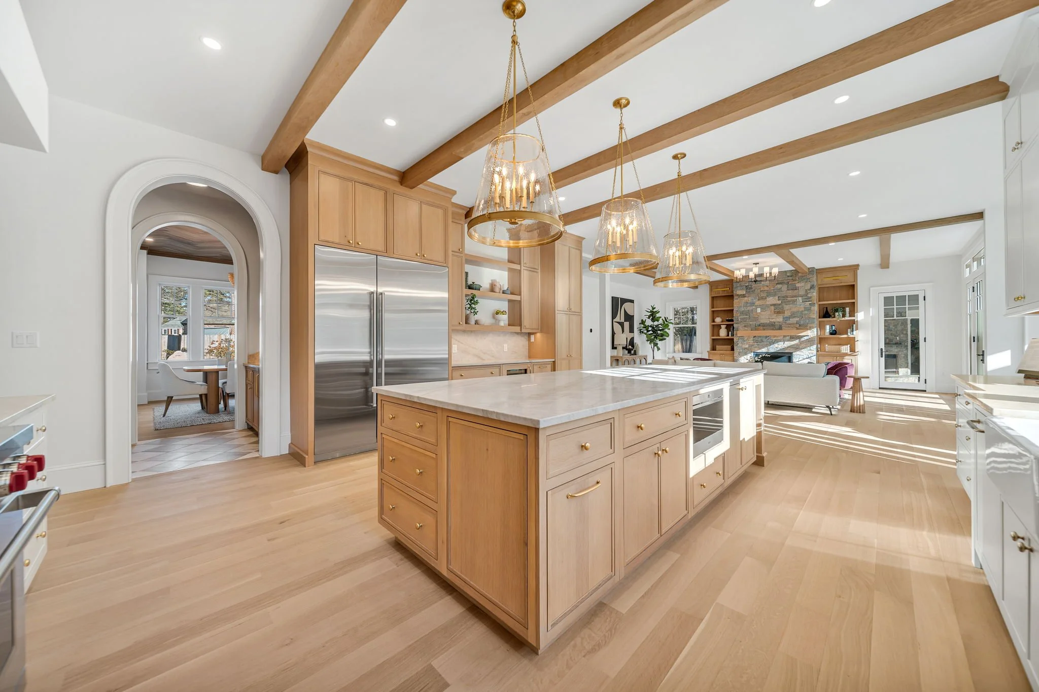 Open-concept kitchen with a large island, wooden cabinets, stainless steel refrigerator, and a living area with a stone fireplace and seating.