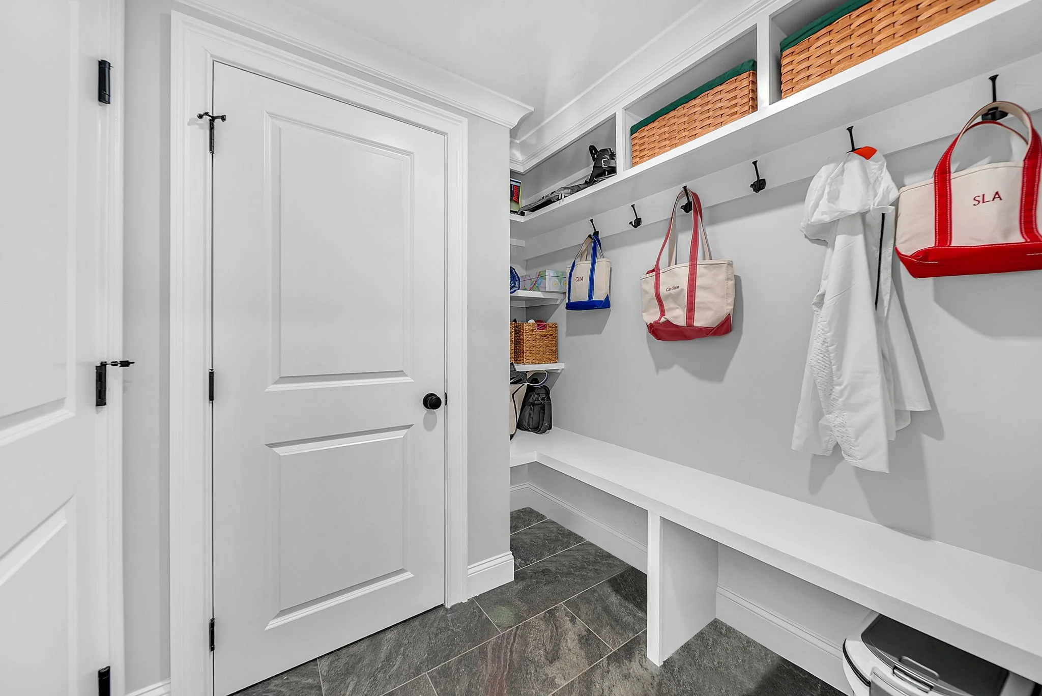 A laundry room with white walls, a white door, black hooks with bags hanging, and white shelves with wicker baskets on top.