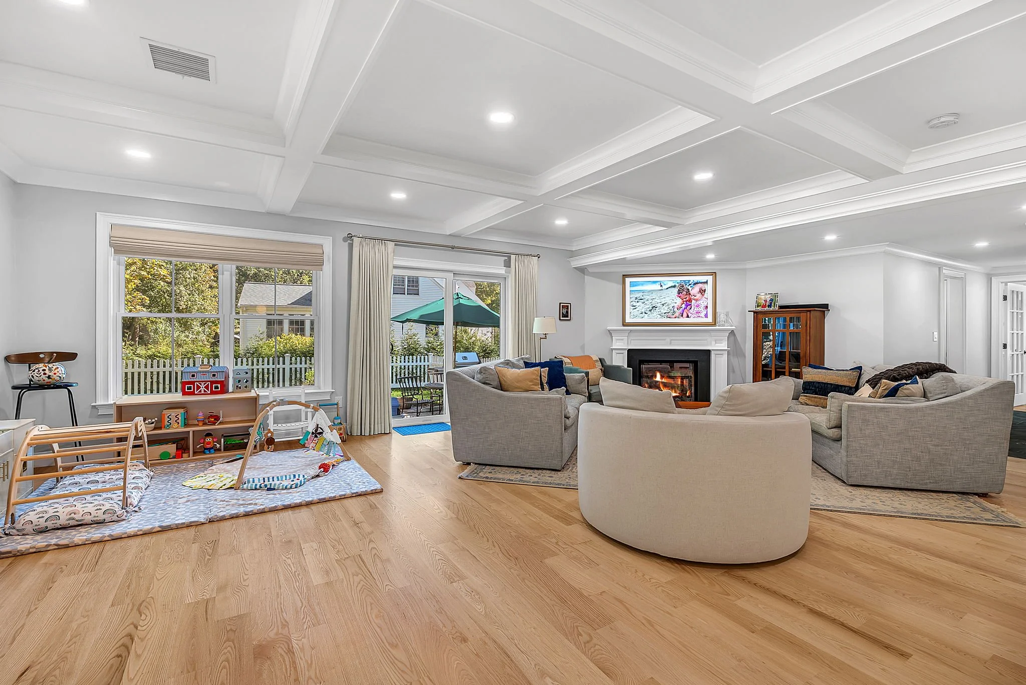 Spacious living room with a cozy fireplace, beige and gray sofas, a wooden cabinet, and large windows with curtains offering a view of an outdoor patio