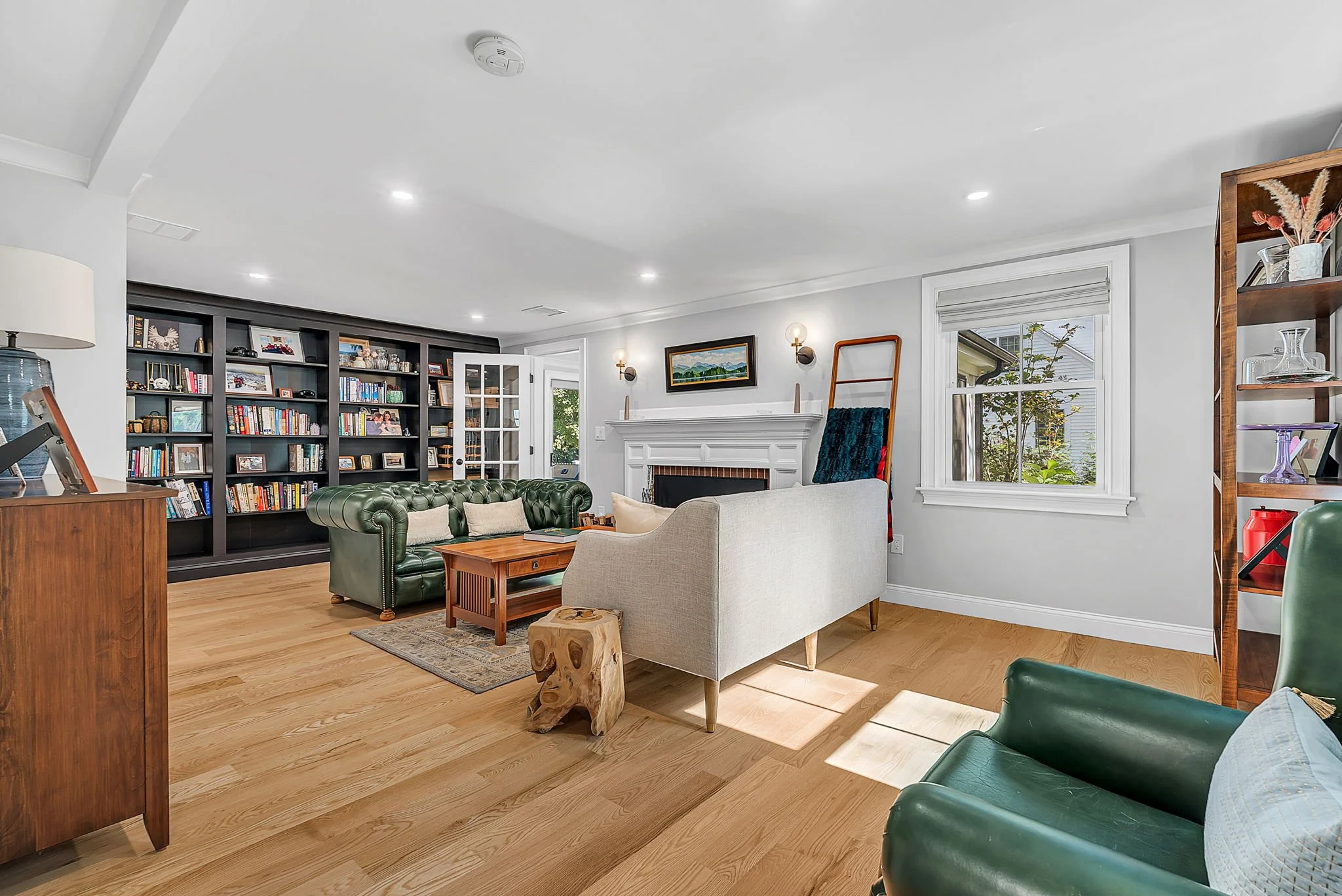 Living room with white walls, hardwood floors, fireplace, black bookshelves filled with books and picture frames, gray and green seating, wooden coffee table, side table, and decorative items, windows with white trim, and a bright, airy atmosphere.