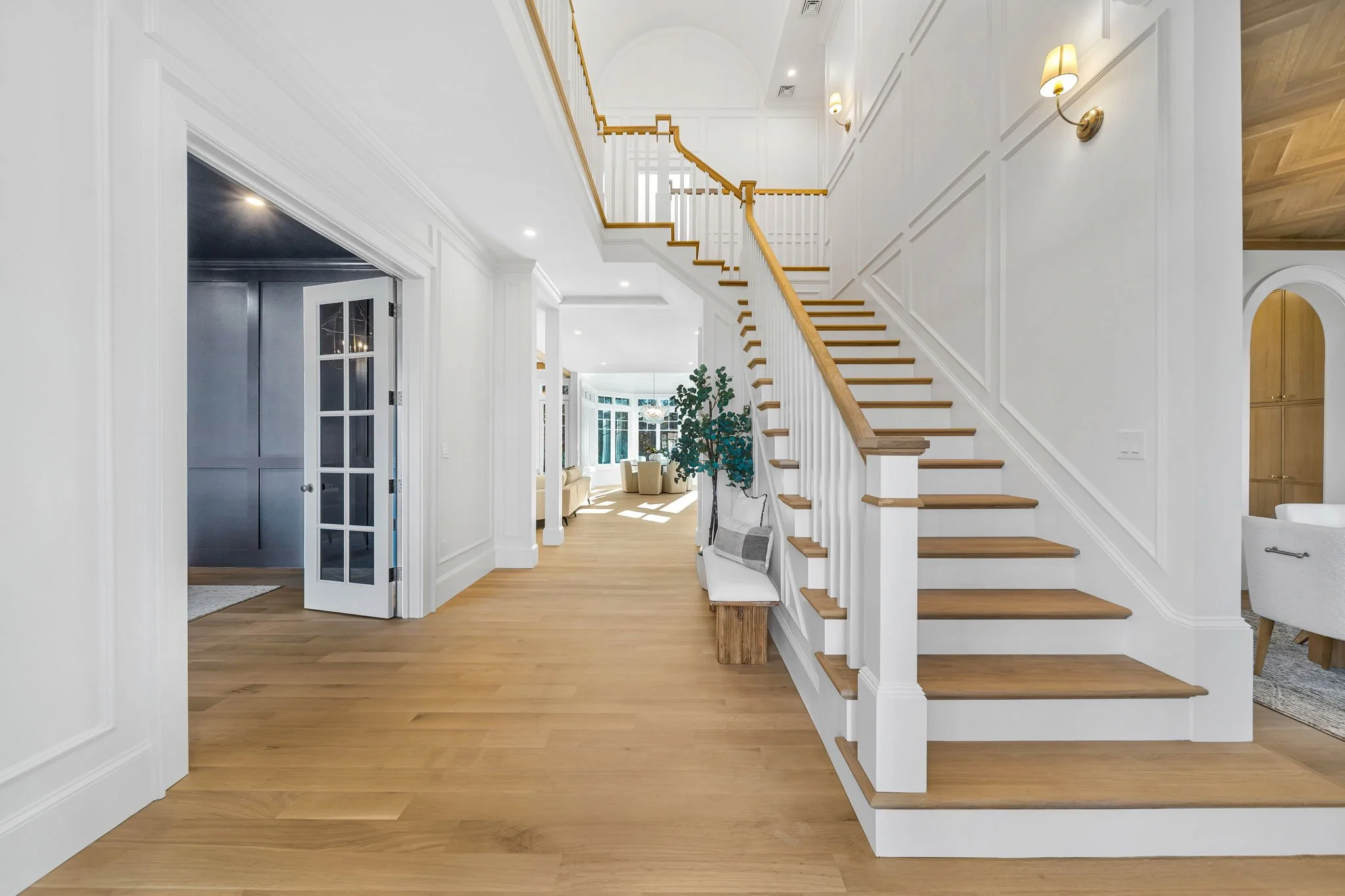 Bright foyer with wooden staircase and white walls, leading into a living room with beige sofas and large windows.