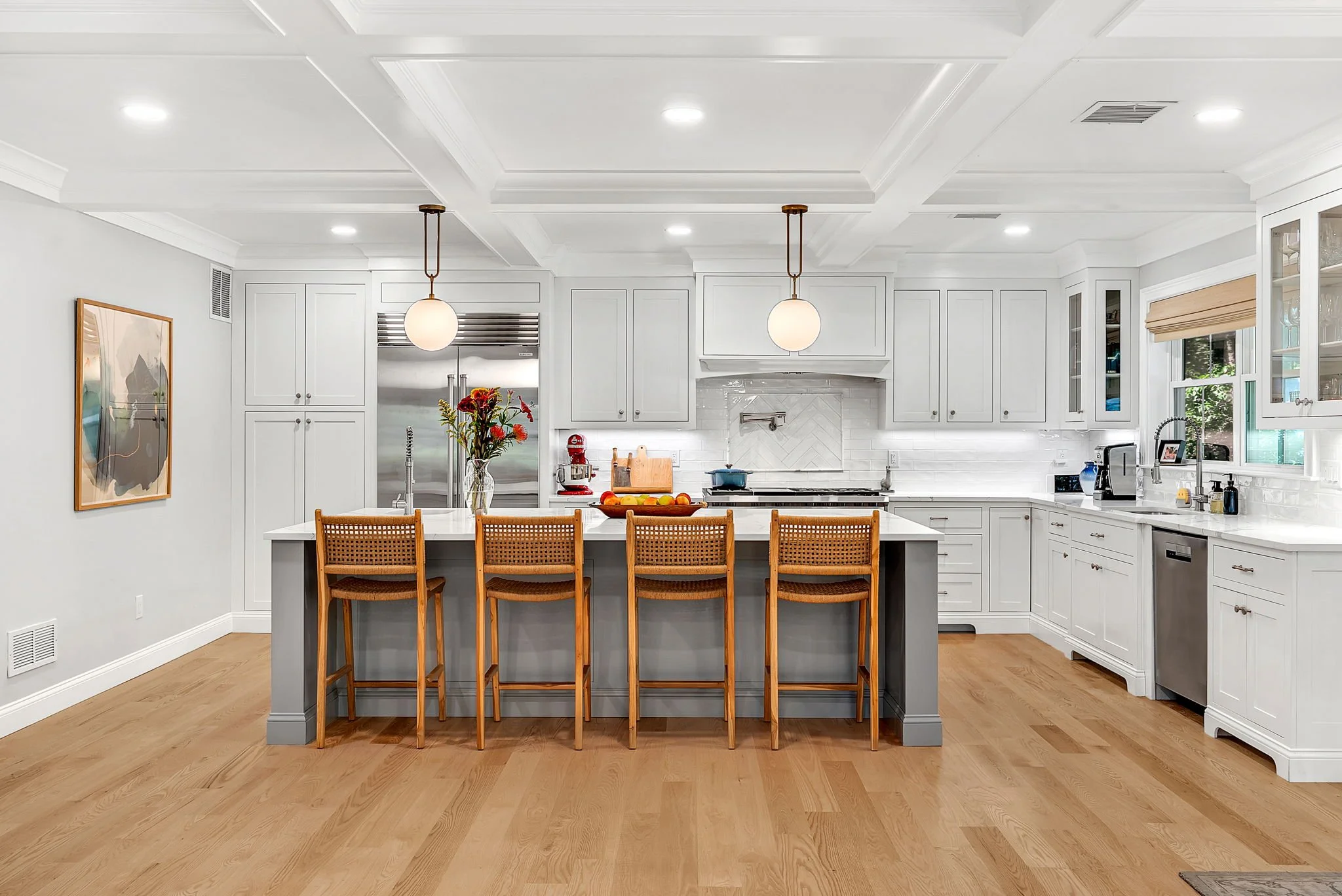 Modern kitchen with white cabinets, island with four wooden bar stools, hardwood floors, stainless steel refrigerator, and large windows.