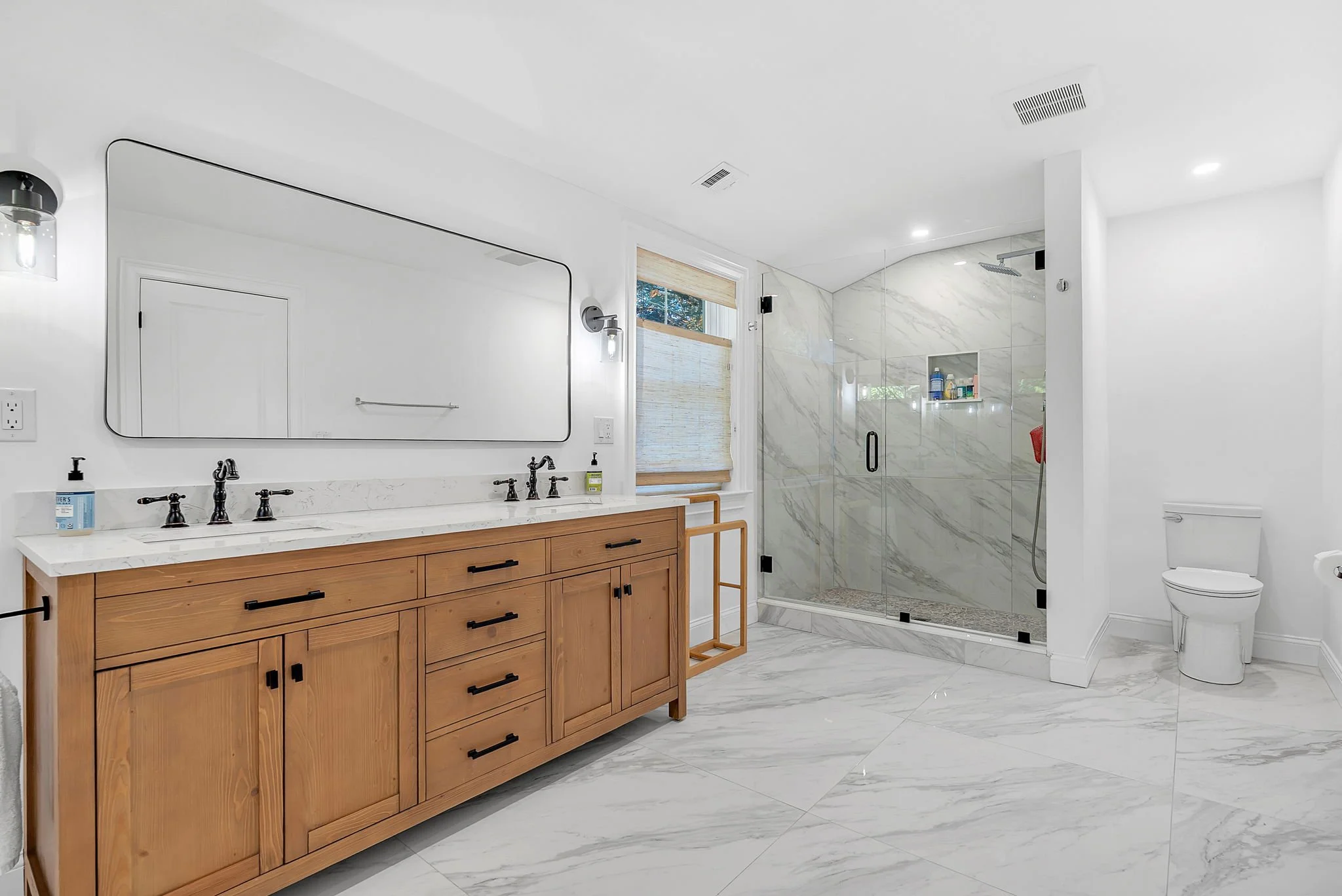 Modern bathroom with wooden vanity, large wall mirror, marble floor and shower with glass door.