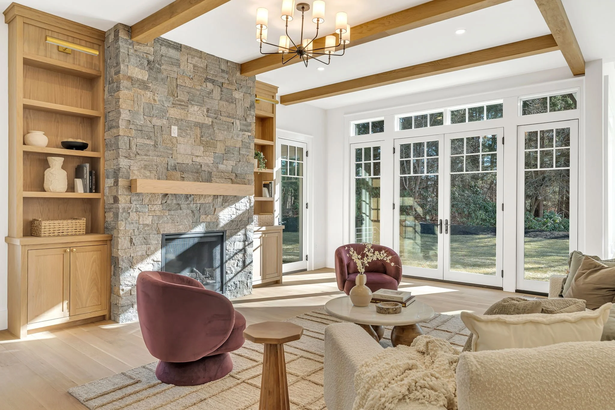 Bright living room with large glass doors, stone fireplace, wooden ceiling beams, built-in bookshelves, and cozy seating including two maroon velvet chairs, a beige sofa with pillows, and a round coffee table with books and a vase.