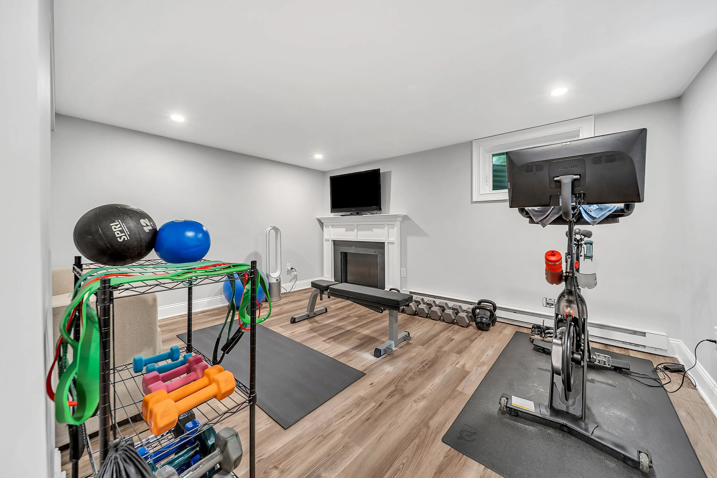 Home gym with exercise equipment including a stationary bike, dumbbells, kettlebells, medicine balls, resistance bands, and a weight bench, with a mounted TV above a fireplace in a room with hardwood flooring and white walls.