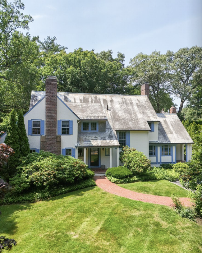 A traditional two-story house with a pitched roof, blue shutters, and brick chimneys, surrounded by green trees and landscaped yard.