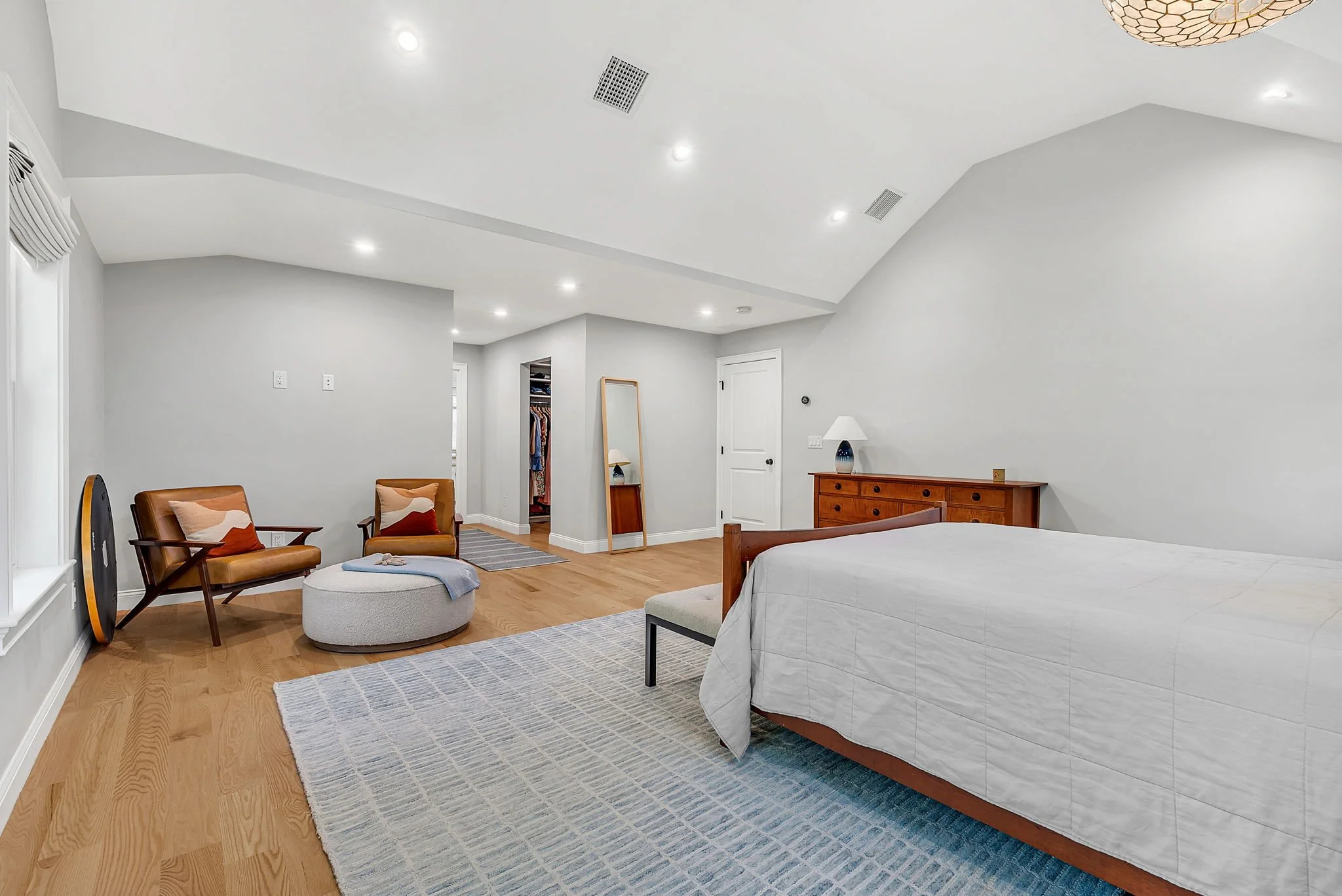 A spacious bedroom with a wooden bed, white bedding, a wooden dresser, a lamp, two armchairs with cushions, an ottoman with a blanket, a mirror, a closet, and hardwood floors.