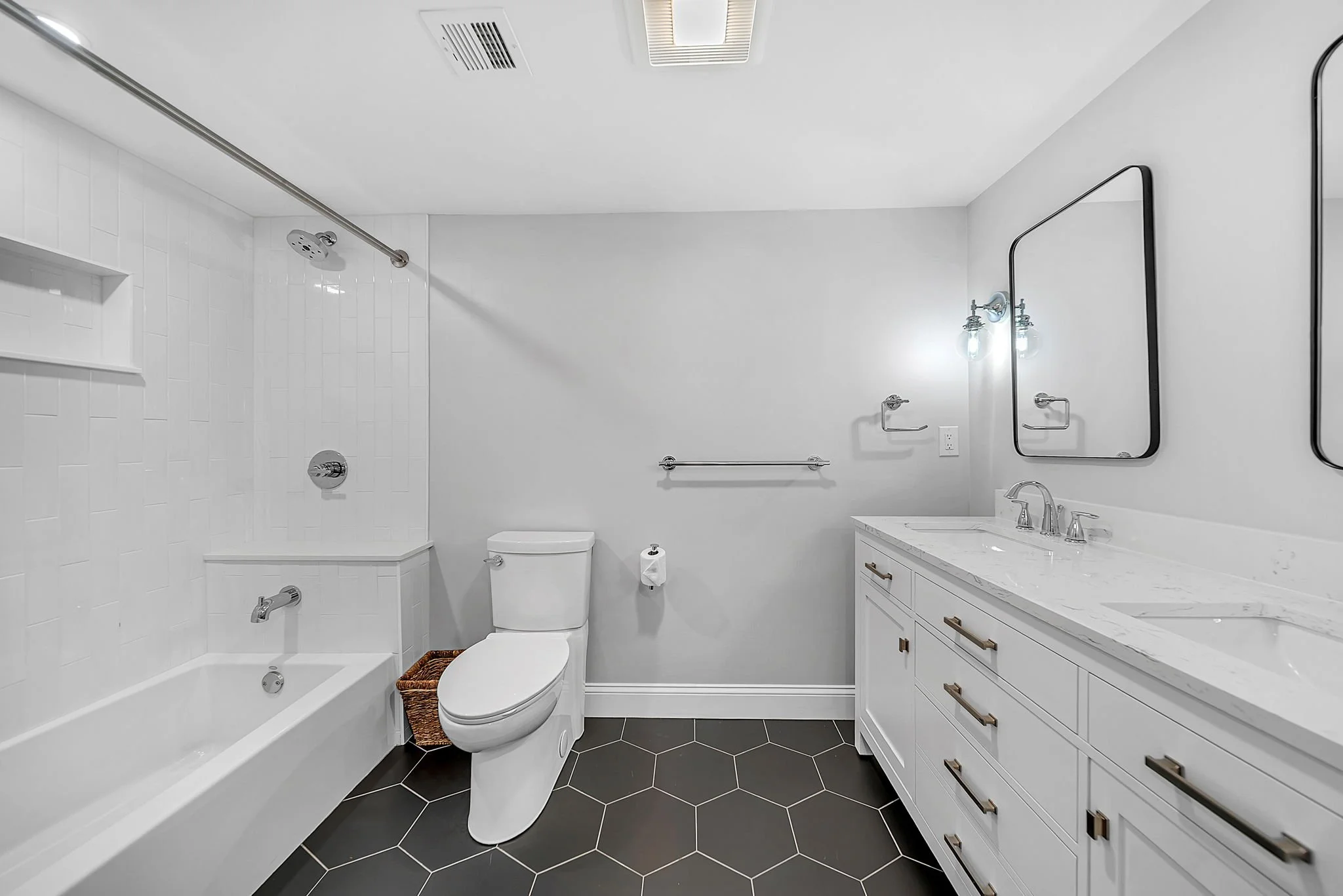 Modern bathroom featuring a white bathtub and shower, toilet, white vanity with a marble countertop, and large mirrors with black frames, black hexagonal floor tiles, and wall-mounted lighting fixtures.