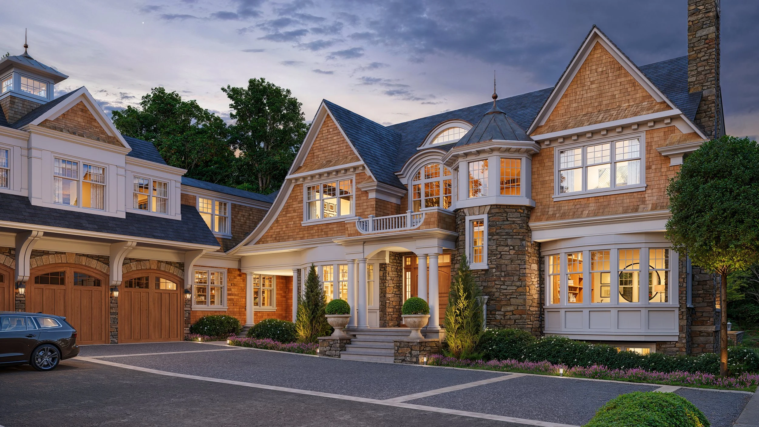 A large luxurious house with multiple levels, stone and wood exterior, large windows, and a front porch with columns. There are trees and landscaped gardens around it, and a parking area with a car in the driveway. The sky is partly cloudy and it appears to be evening.