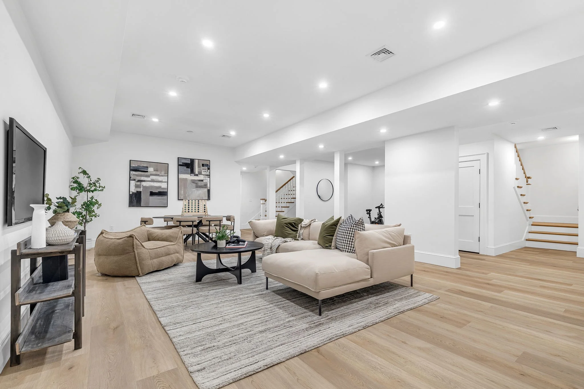 Spacious modern living room with beige sectional sofa, wooden flooring, black coffee table, round mirror, artwork, TV, potted plants, and staircase in the background.