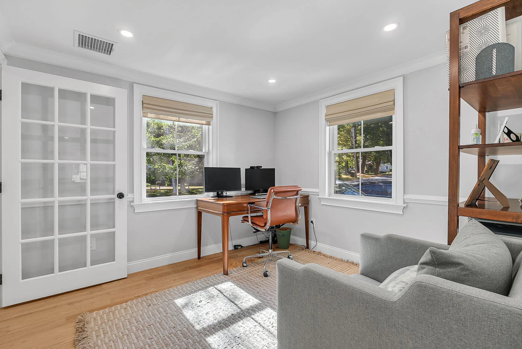 Home office with two computer monitors on a wooden desk, a brown leather chair, beige window shades, white walls, a gray sofa, and a wooden bookshelf.