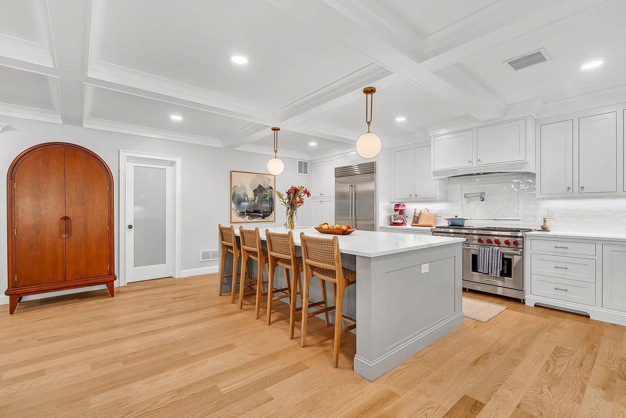 Modern kitchen with white cabinets, stainless steel appliances, light wood flooring, and a large island with four wooden chairs.