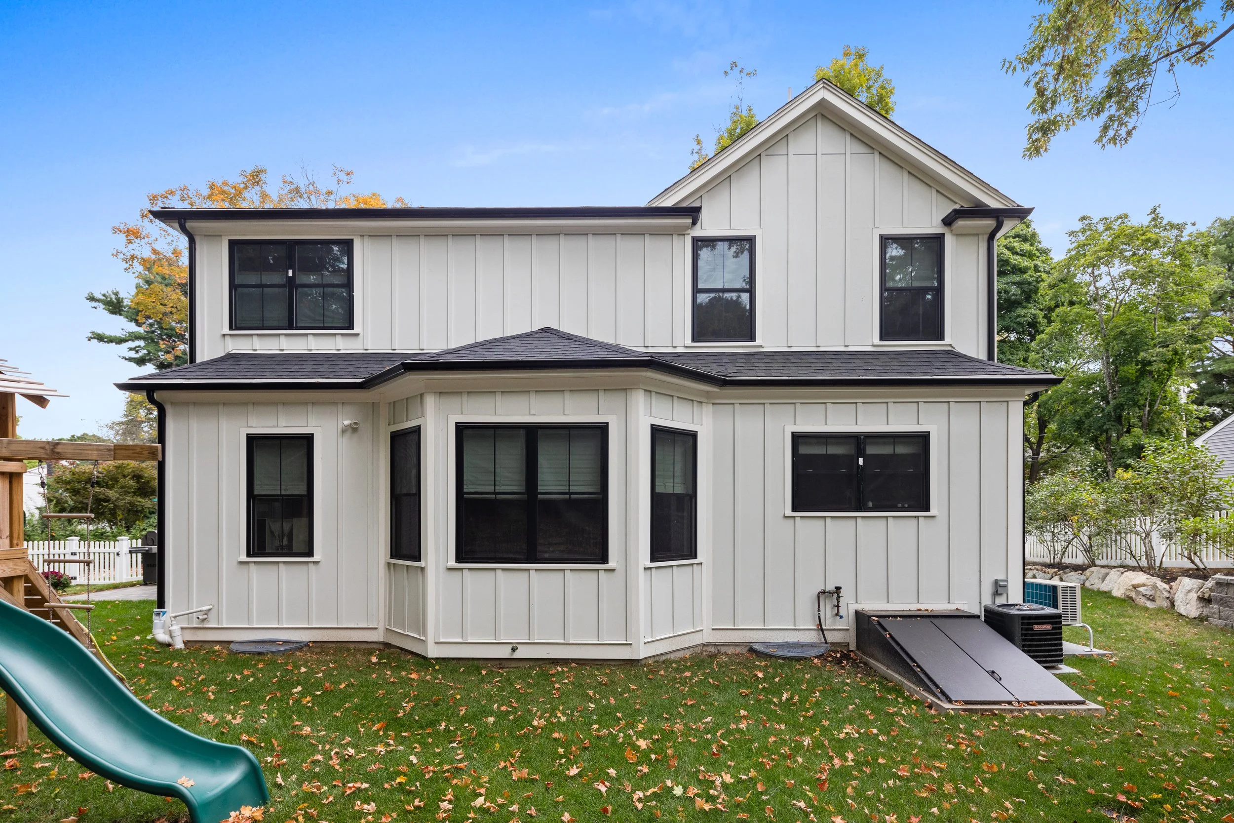 Back view of a two-story white house with black window frames, a black pitched roof, and a small outdoor slide on the grassy yard.