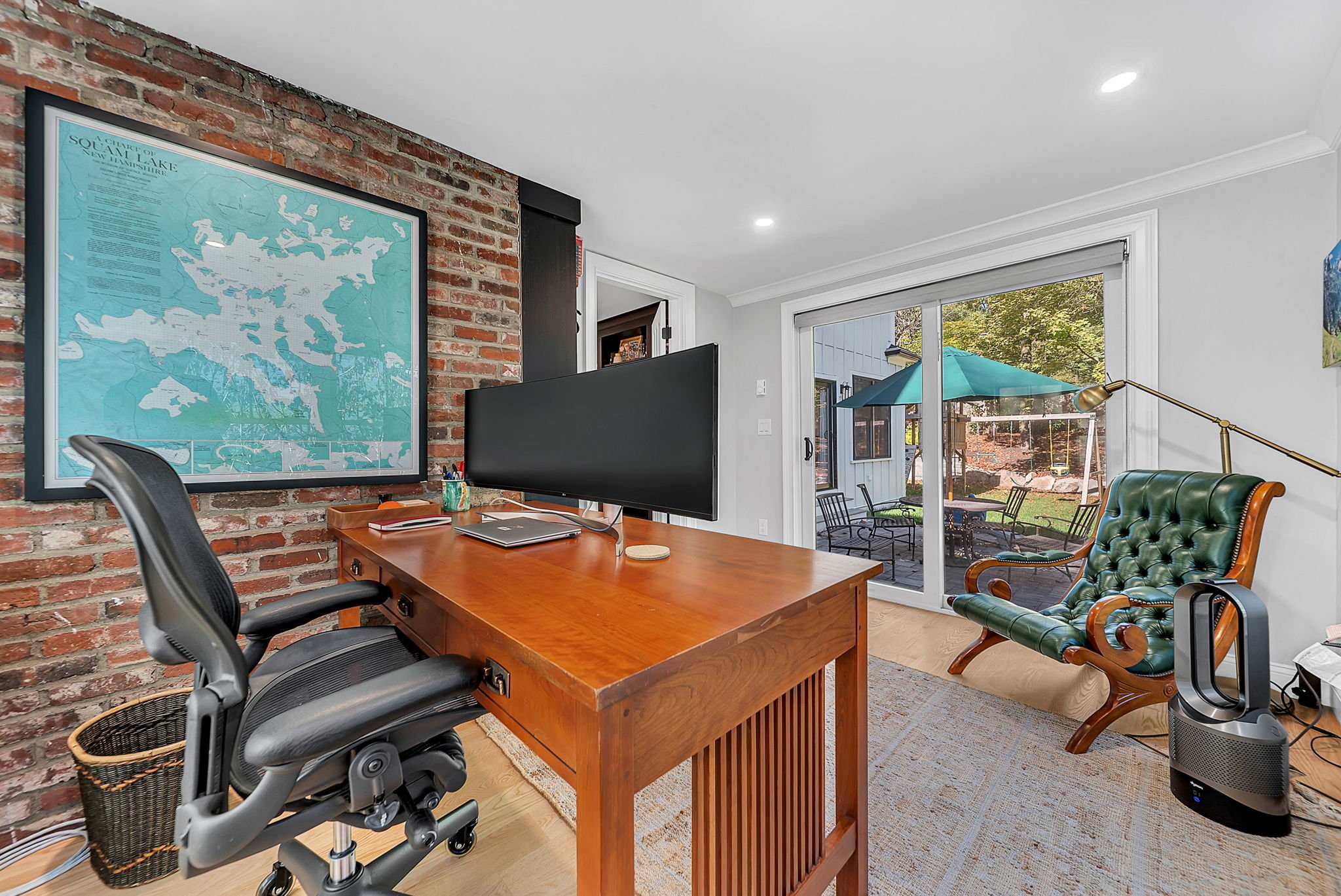 Home office with wooden desk, ergonomic black office chair, large monitor, and wall map, with sliding glass door leading to outdoor patio with table, chairs, and umbrella.