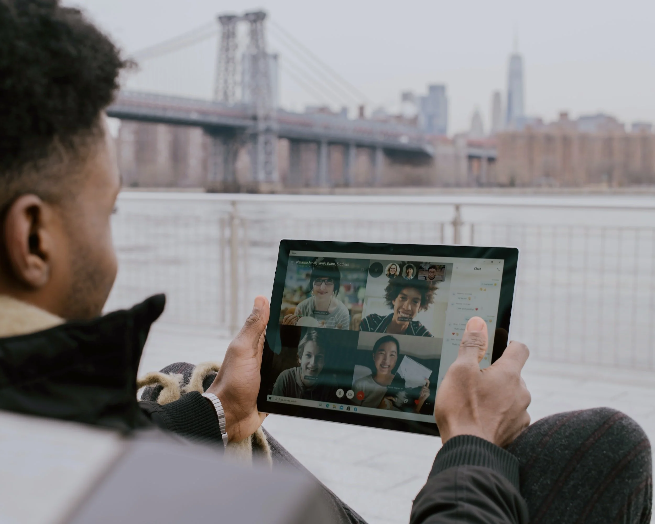 A person sitting outdoors near water, holding a tablet with a video chat with four people, with city buildings and a bridge in the background.