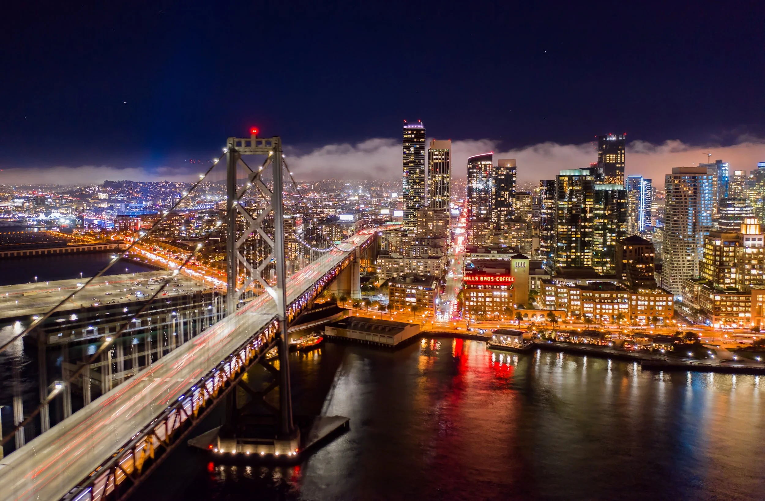 Night view of San Francisco skyline with illuminated city buildings, Bay Bridge with light trails from moving vehicles, and water reflecting city lights.