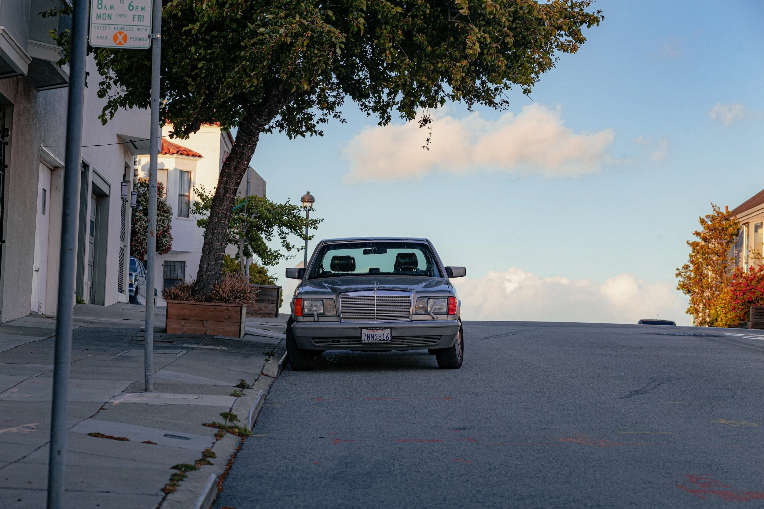Silver Mercedes-Benz sedan parked on the street facing front, with houses and trees on either side of the road, and a partly cloudy sky overhead.