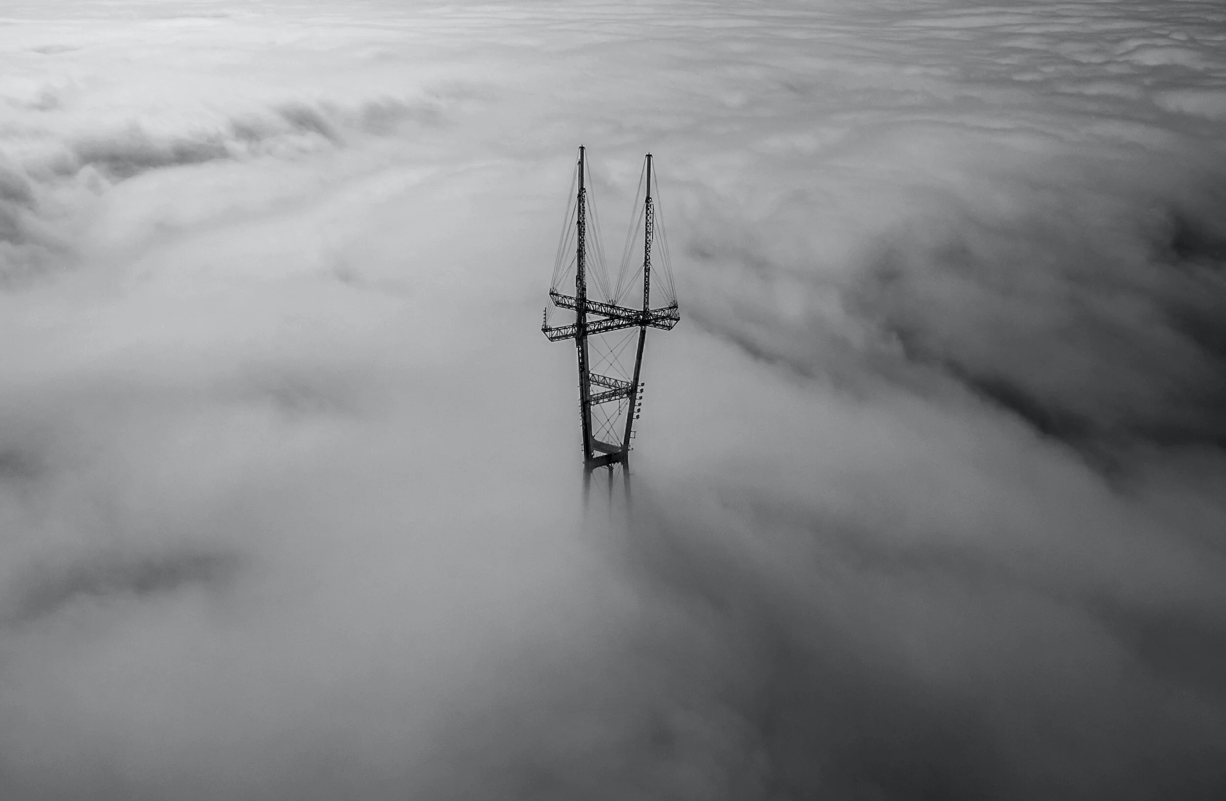 A tall communication tower supported by cables, emerging through a thick layer of clouds or fog.