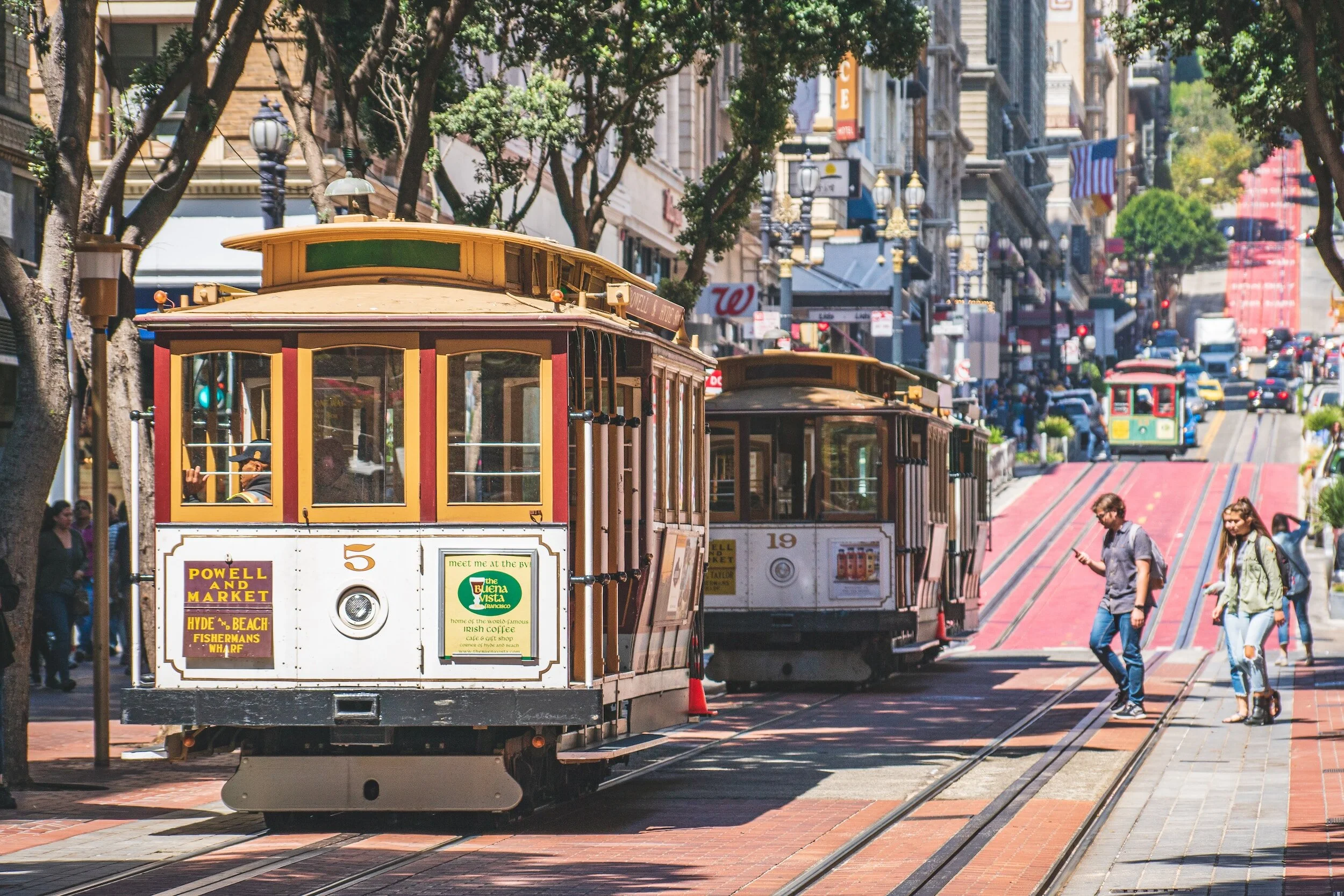 Two vintage cable cars on a city street with people walking nearby