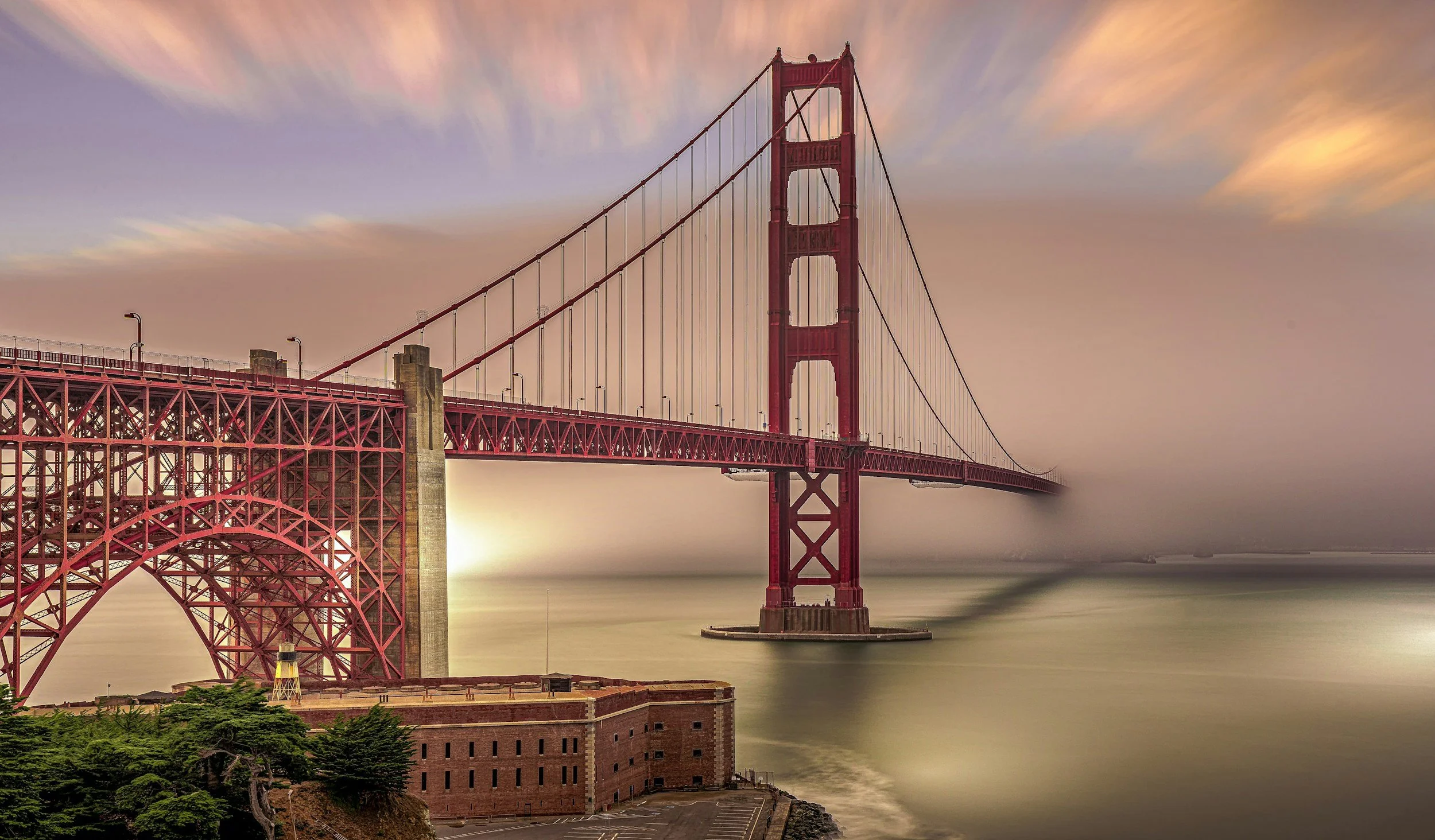 Long exposure photo of the Golden Gate Bridge at sunset, showing its red towers and suspension cables, with a soft sky and water below.