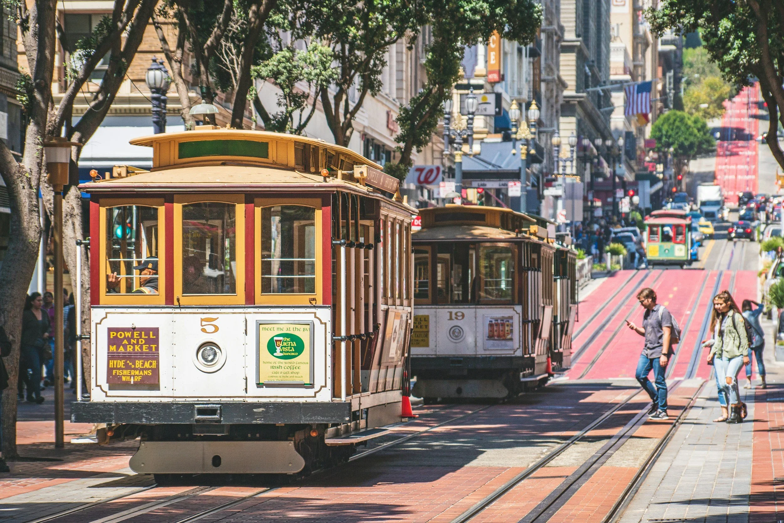 Two vintage streetcars on a city street with pedestrians, trees, and buildings in the background.