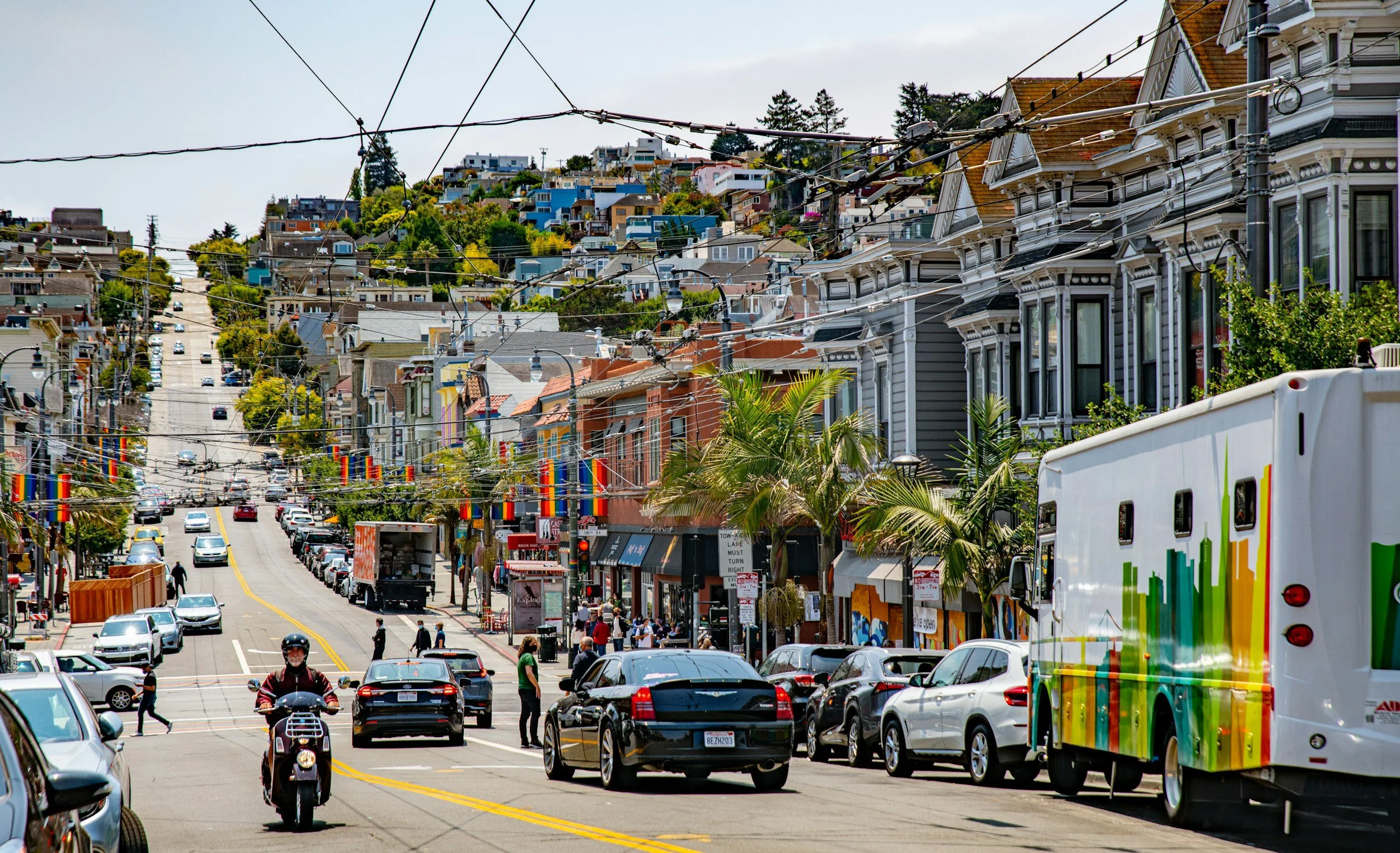 A lively city street with parked cars, pedestrians, colorful banners, and houses on a hillside in the background.