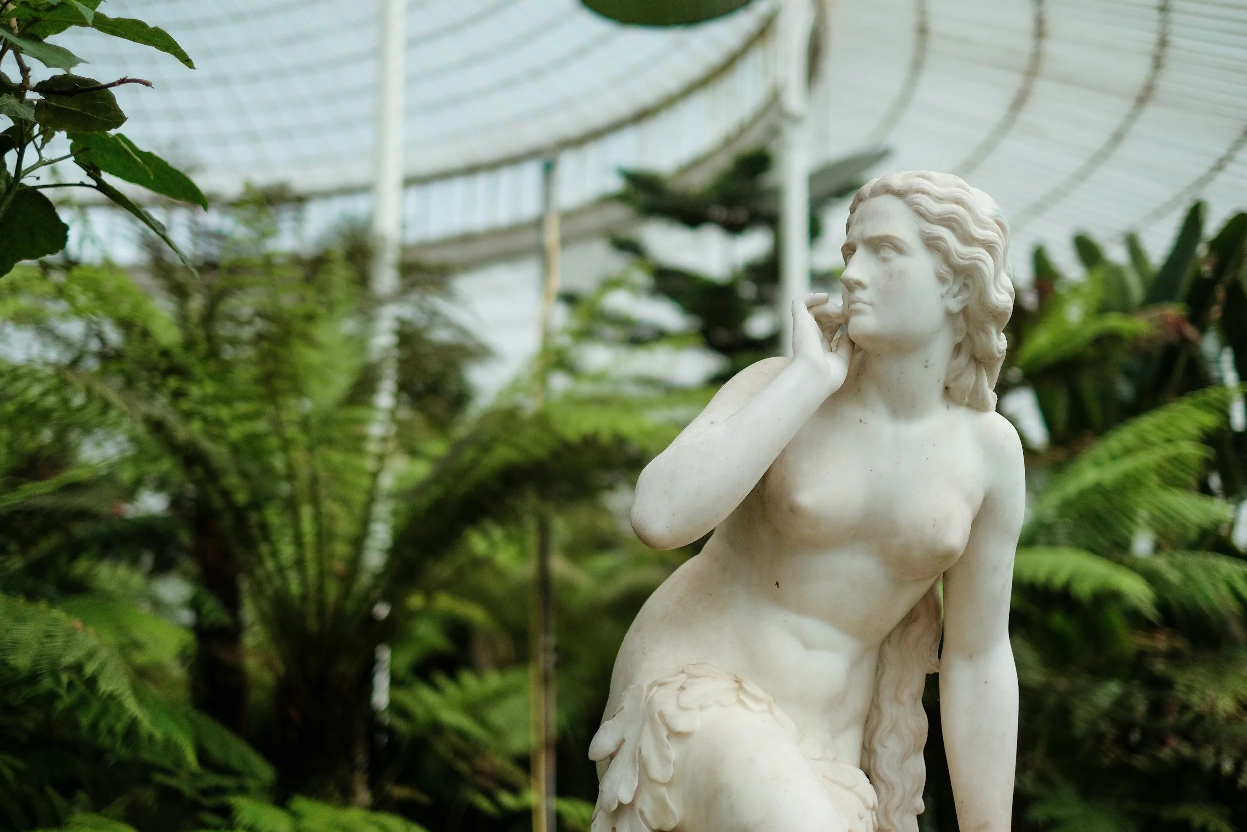 Marble sculpture of a woman in a greenhouse surrounded by lush green plants.